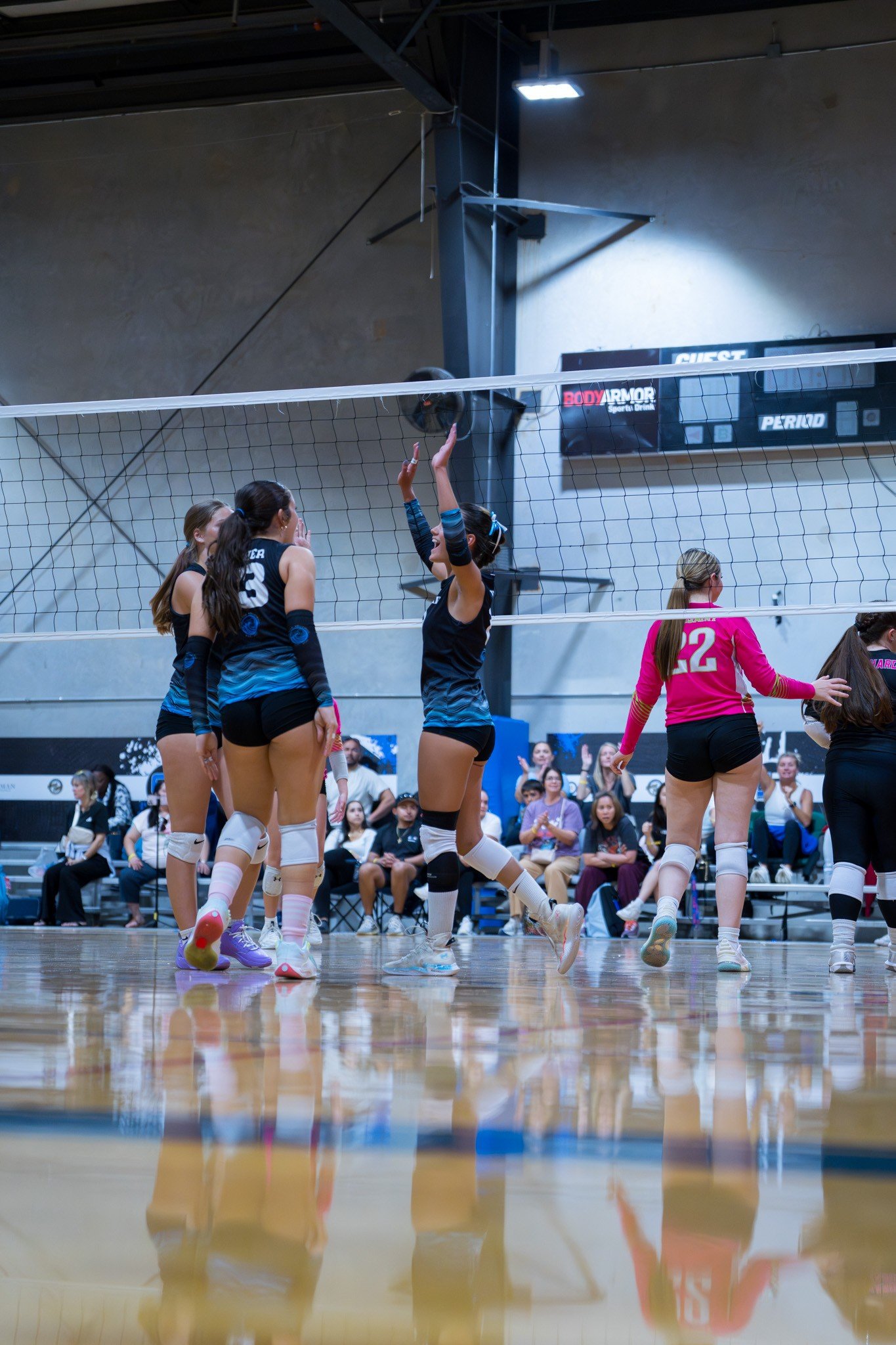 Girls playing volleyball in an indoor gymnasium, with one girl in black uniform jumping near the net while her teammates and opponents watch.