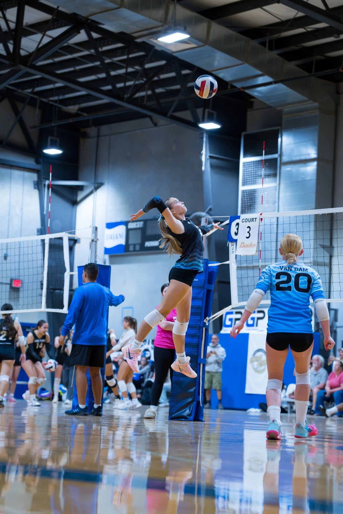 A female volleyball player in black uniform jumping to hit the ball over the net during a game at an indoor volleyball court. Other players and spectators are visible in the background.