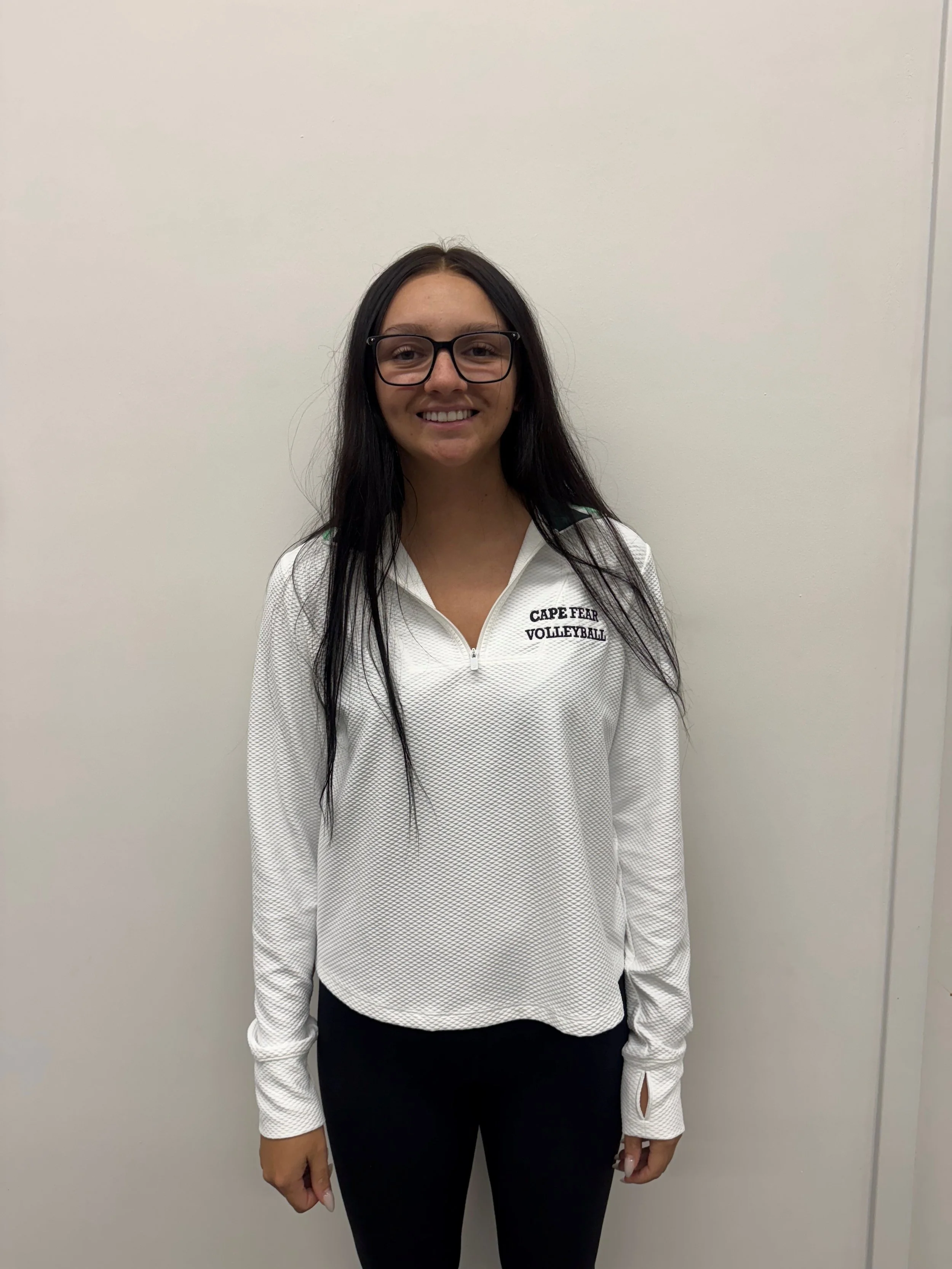 A smiling young woman with long brown hair wearing a maroon sweatshirt with yellow and white text that reads "Concordia University Chicago". She is standing in front of a textured light yellow wall with a decorative metal sun and plant wall hanging.