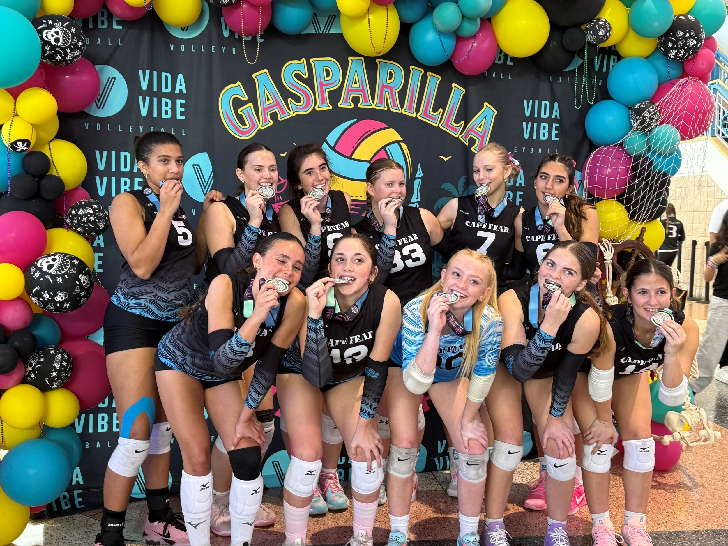 A group of young women volleyball players in black uniforms are celebrating their victory, biting medals, and posing in front of a colorful GaspArilla backdrop with balloons. They are wearing knee pads and sports gear.