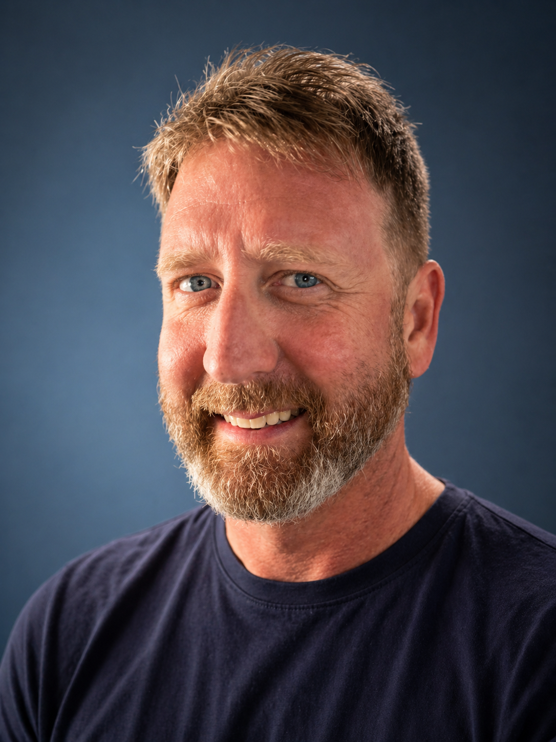 Close-up of a smiling middle-aged man with a beard and blue eyes, wearing a black t-shirt, against a solid dark blue background.