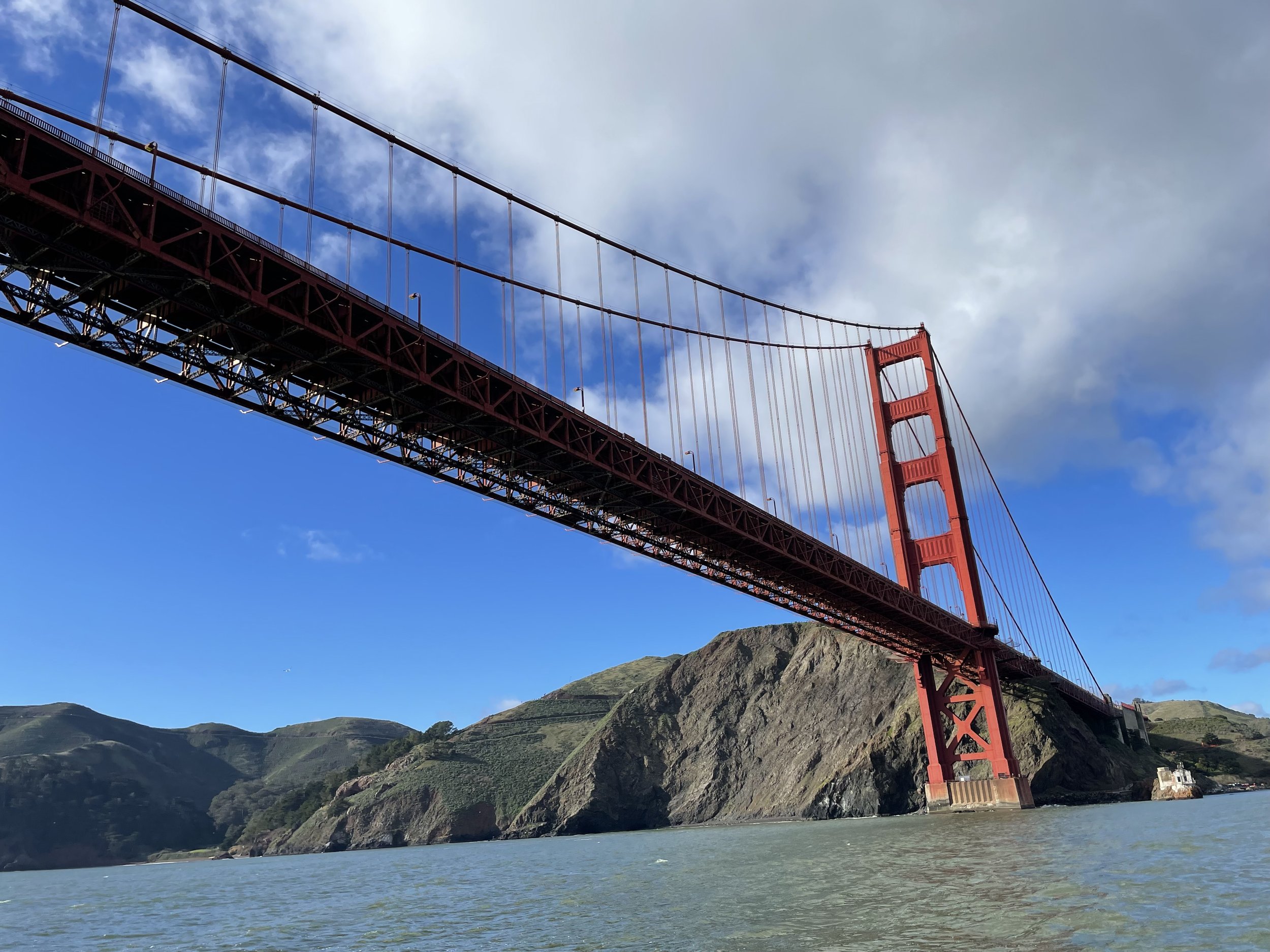 View of the Golden Gate Bridge in San Francisco with clear blue sky, some clouds, and green hills in the background.