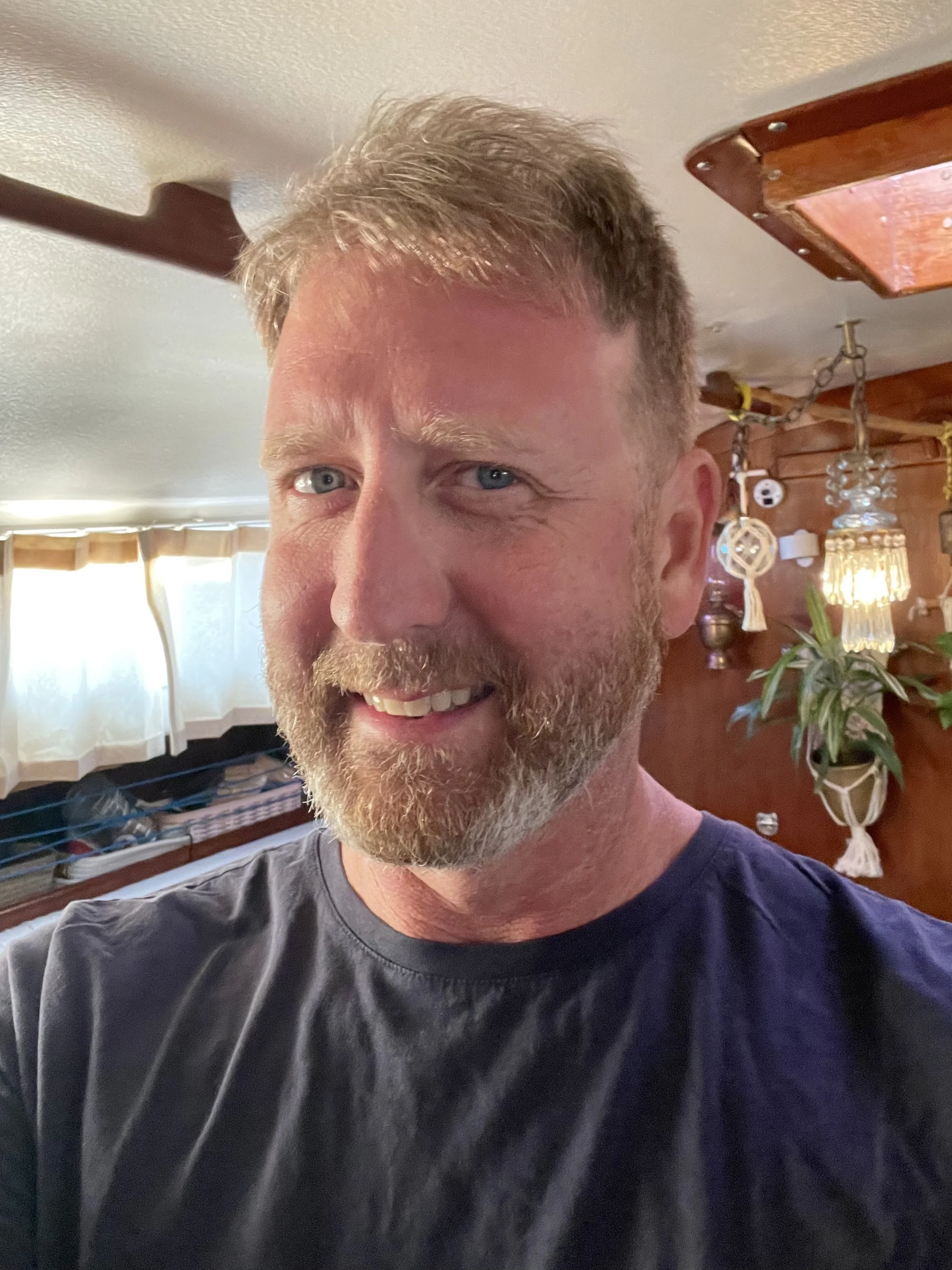 A man with a beard and short hair smiling, taking a selfie in a room with wooden walls, plants, and decorative hanging lights.