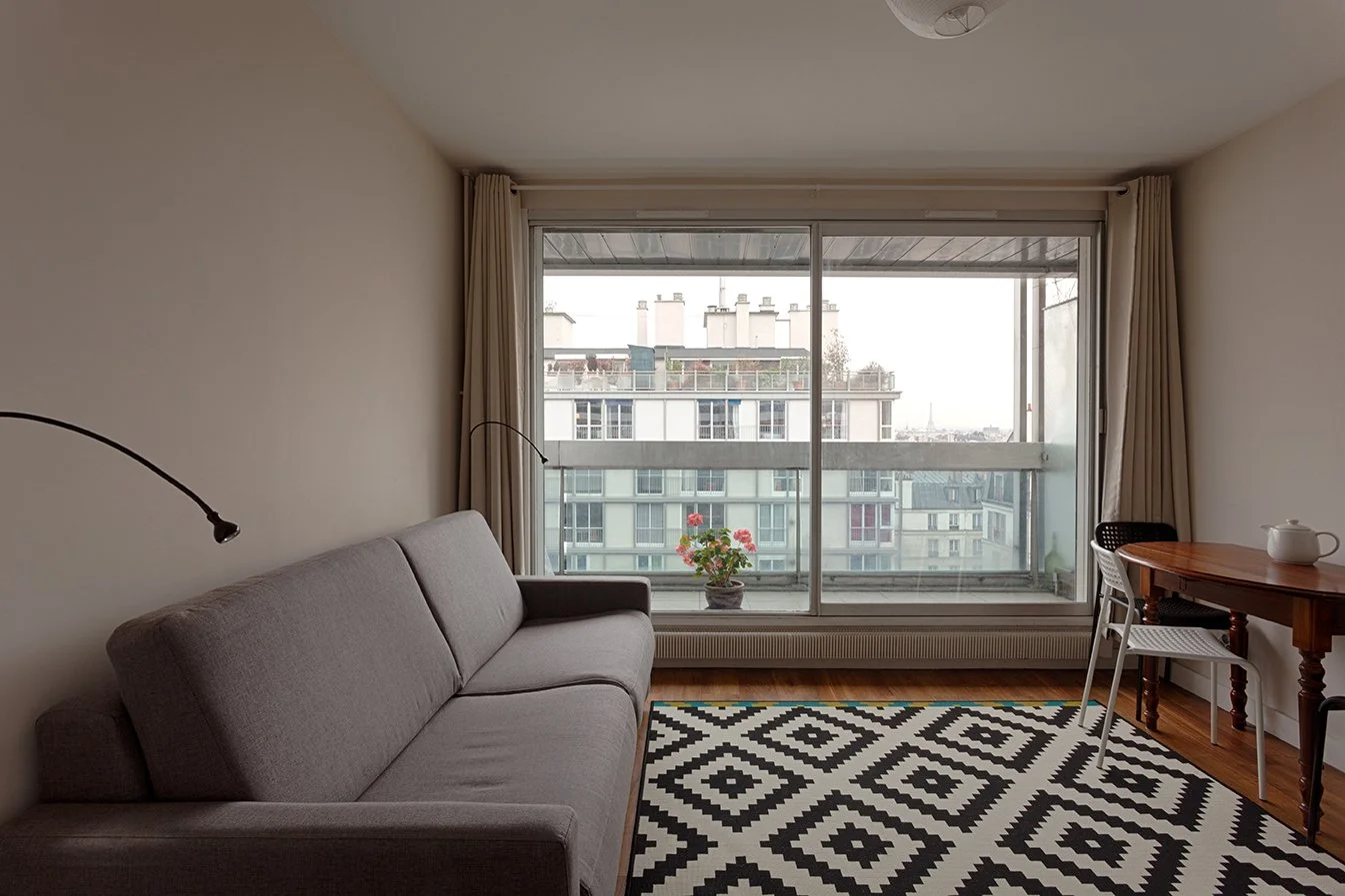 Salon avec un canapé gris, une table en bois avec une théière, une plante en pot sur le balcon, grands rideaux blancs, tapis noir et blanc à motifs, vue sur des immeubles de l'extérieur