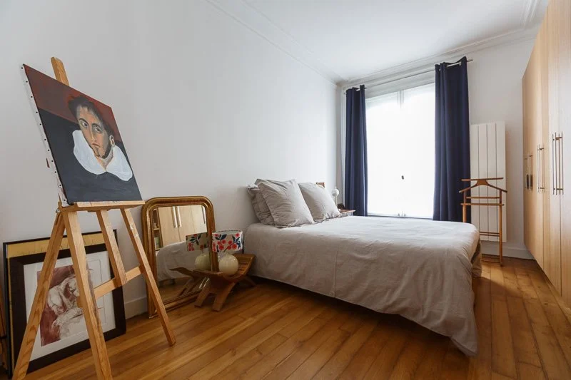 Chambre simple avec un lit en bois, des oreillers, des œuvres d'art sur un chevalet, et une grande fenêtre avec rideaux bleu foncé.