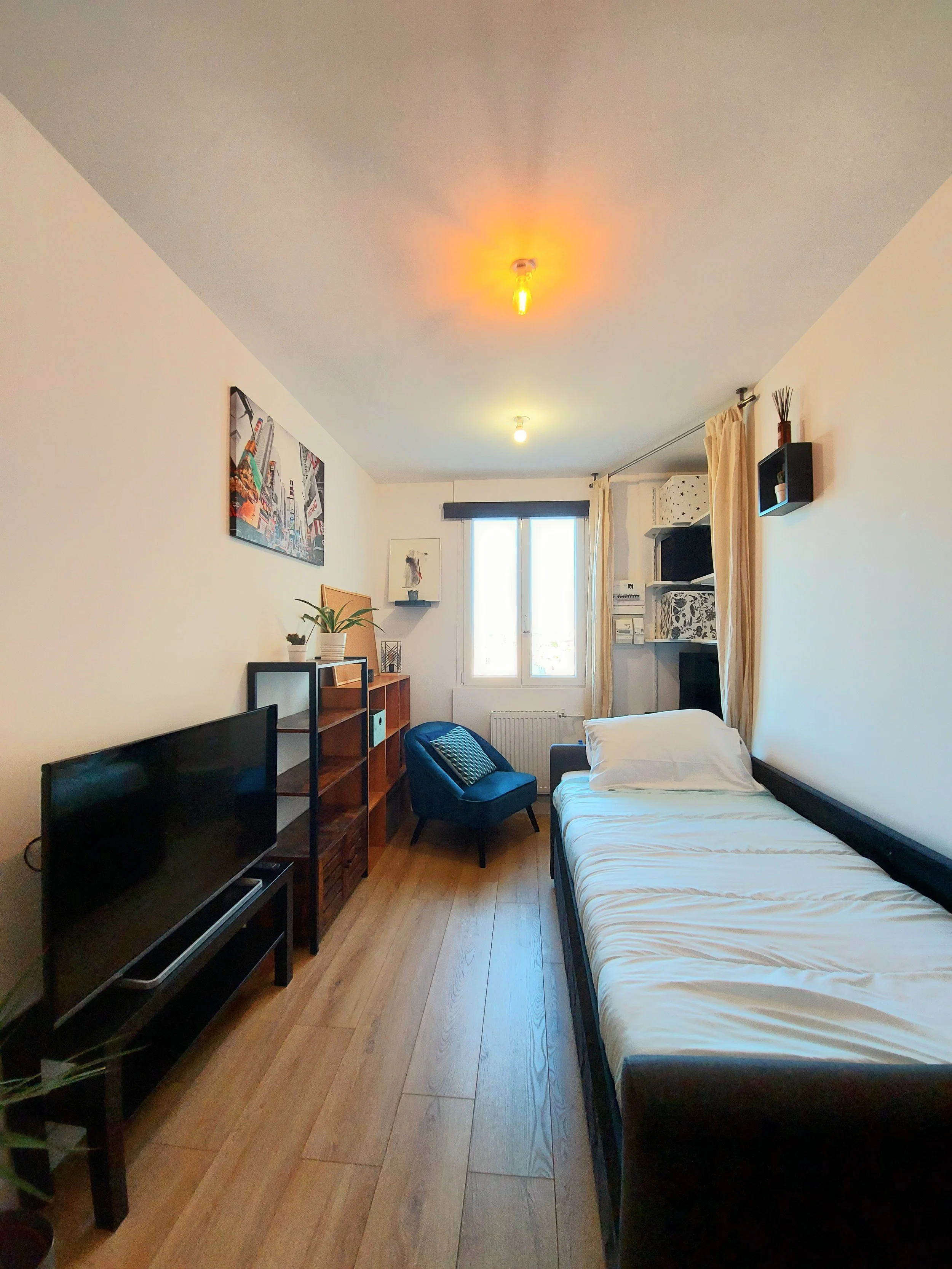 Chambre avec un lit simple, un fauteuil bleu, une télévision, une étagère en bois, une fenêtre avec rideaux beiges, un mur décoré d'une affiche artistique, et plusieurs petites décorations.
