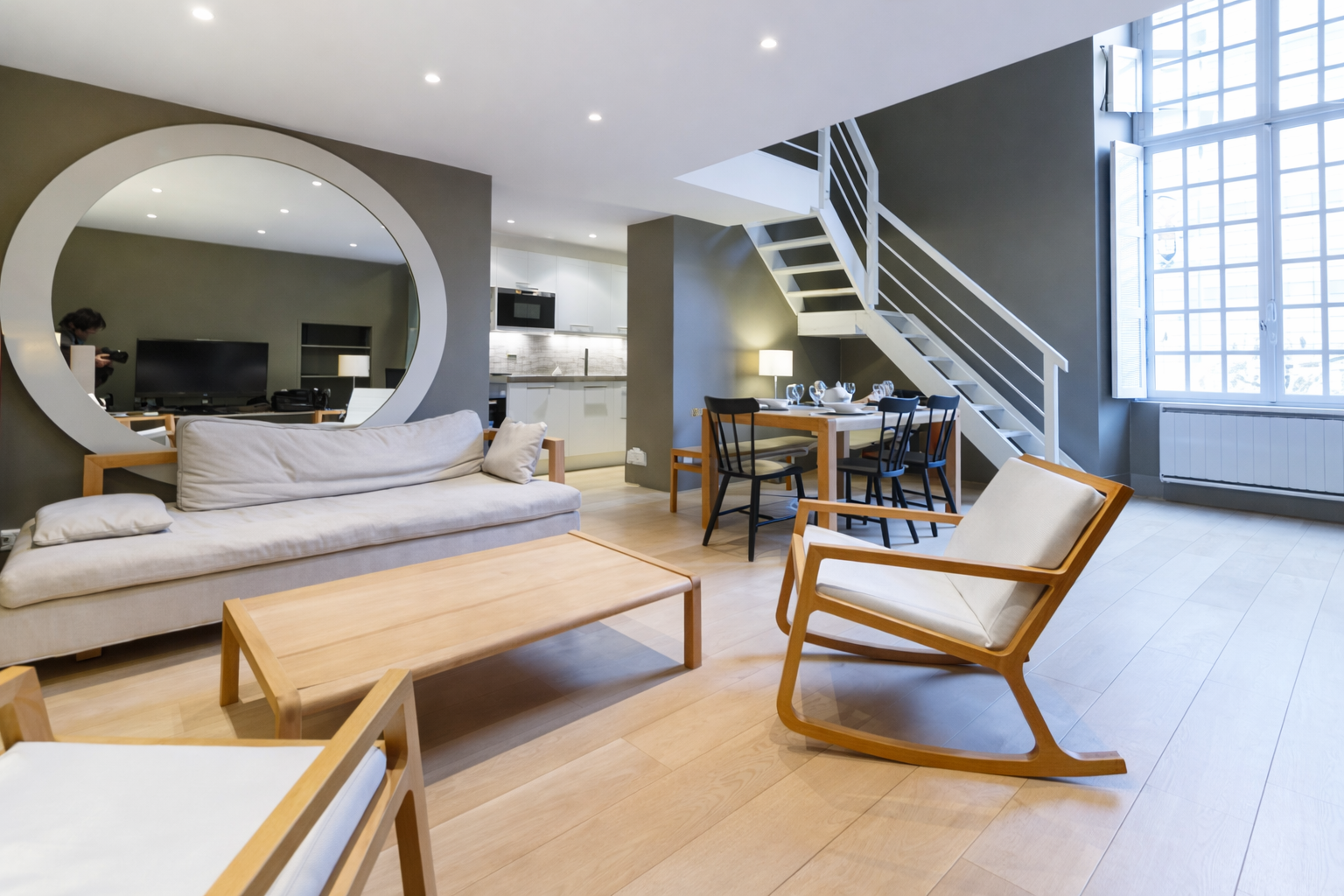 Intérieur moderne d'un salon et coin repas avec canapé, fauteuil en bois, table basse, grande fenêtre, escalier blanc, et cuisine en arrière-plan.