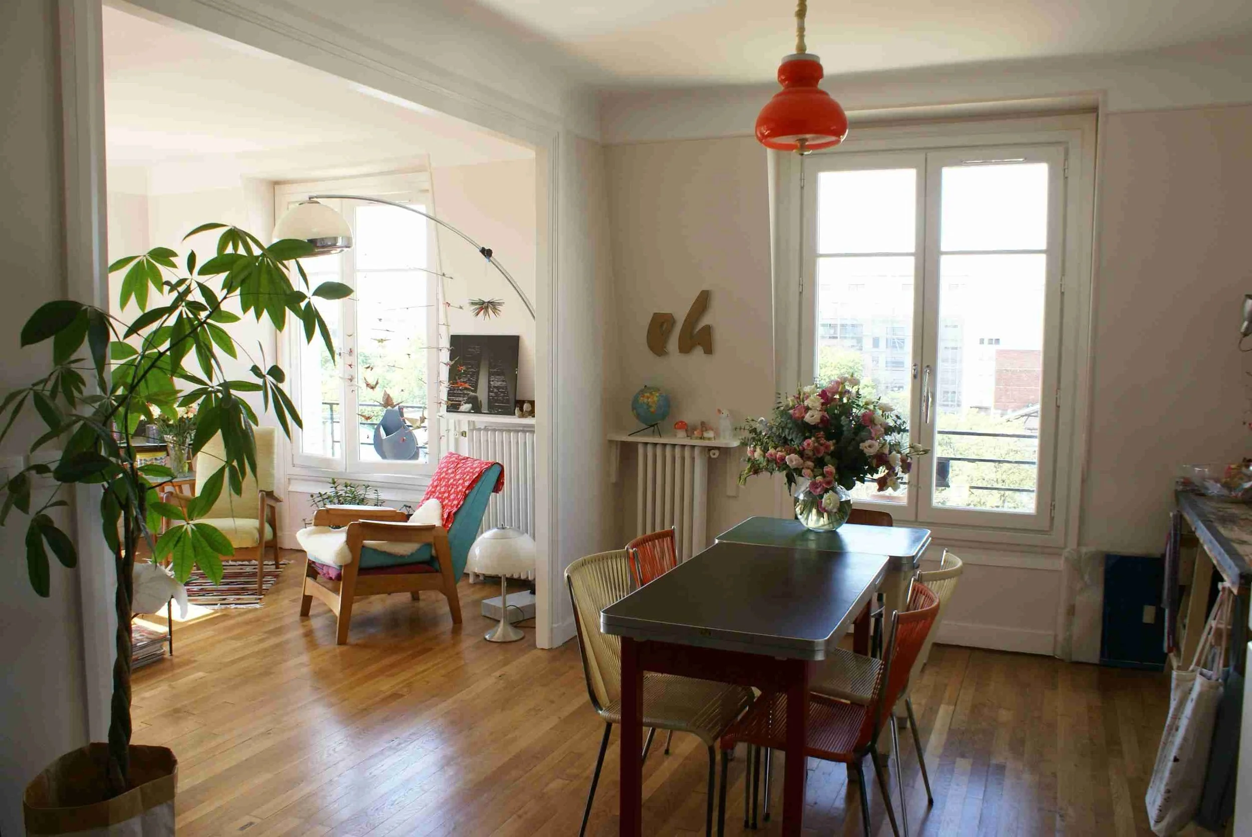 Une salle à manger lumineuse avec une table entourée de chaises, un vase avec des fleurs sur la table, et une fenêtre laissant entrer la lumière naturelle. Un coin salon avec des fauteuils et des décorations.