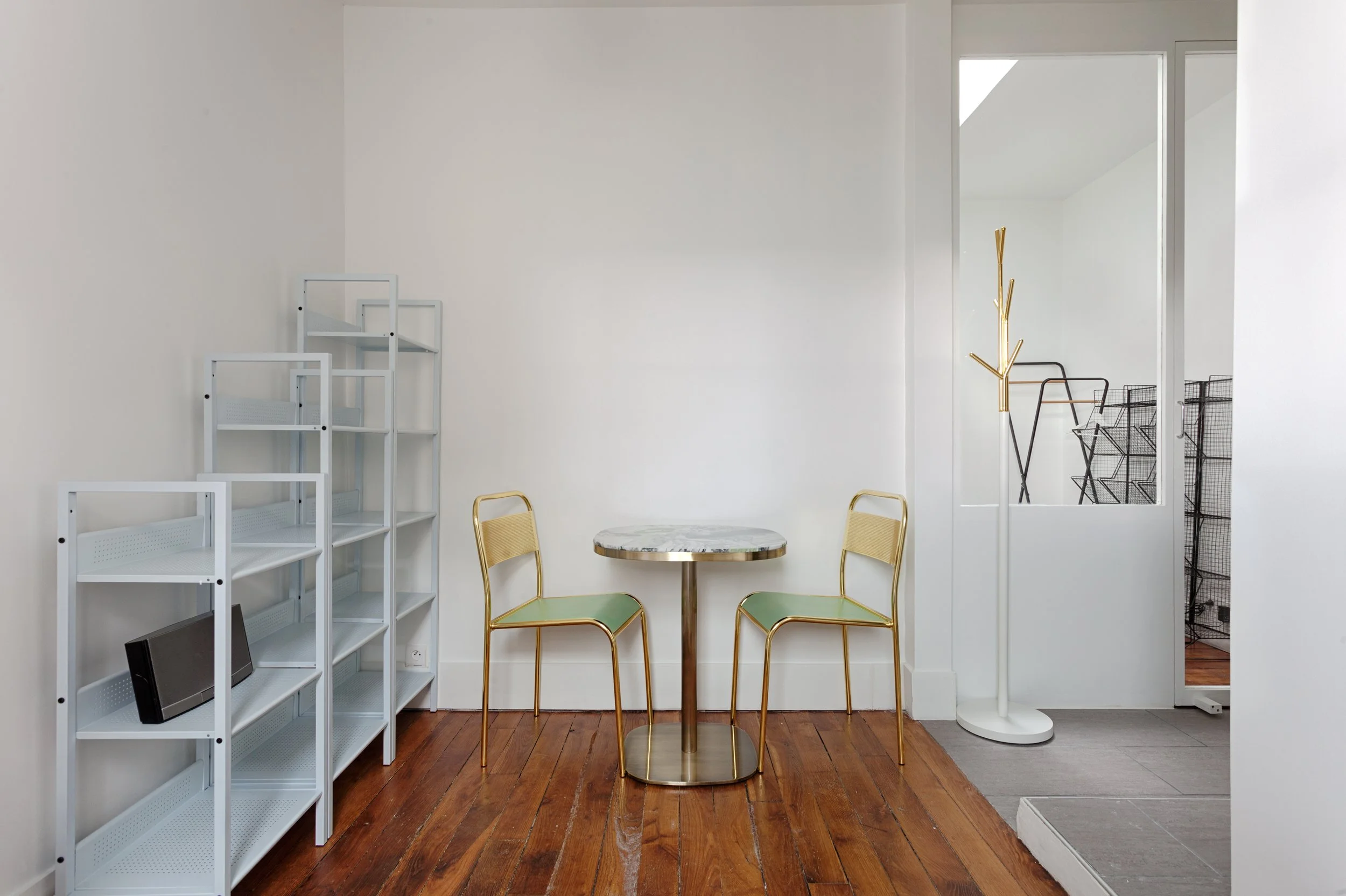 Une pièce minimaliste avec un sol en bois, un mur blanc, deux chaises avec cadre en métal doré et assise en vert, une petite table ronde avec un dessus en marbre, des étagères blanches vides, et un porte-manteaux en forme d'arbre en métal doré, une zone de transition avec un sol en carrelage gris et une bannière blanche.