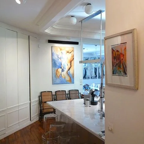 Salon avec une table en marbre, plusieurs fauteuils en bois, œuvres d'art accrochées au mur et une cuisine ouverte en arrière-plan.