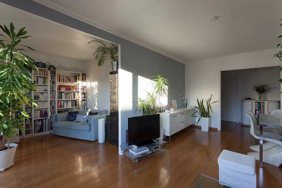 Salon intérieur avec plantes vertes, canapé gris, étagères remplies de livres, télévision, meuble blanc, table à manger avec chaises