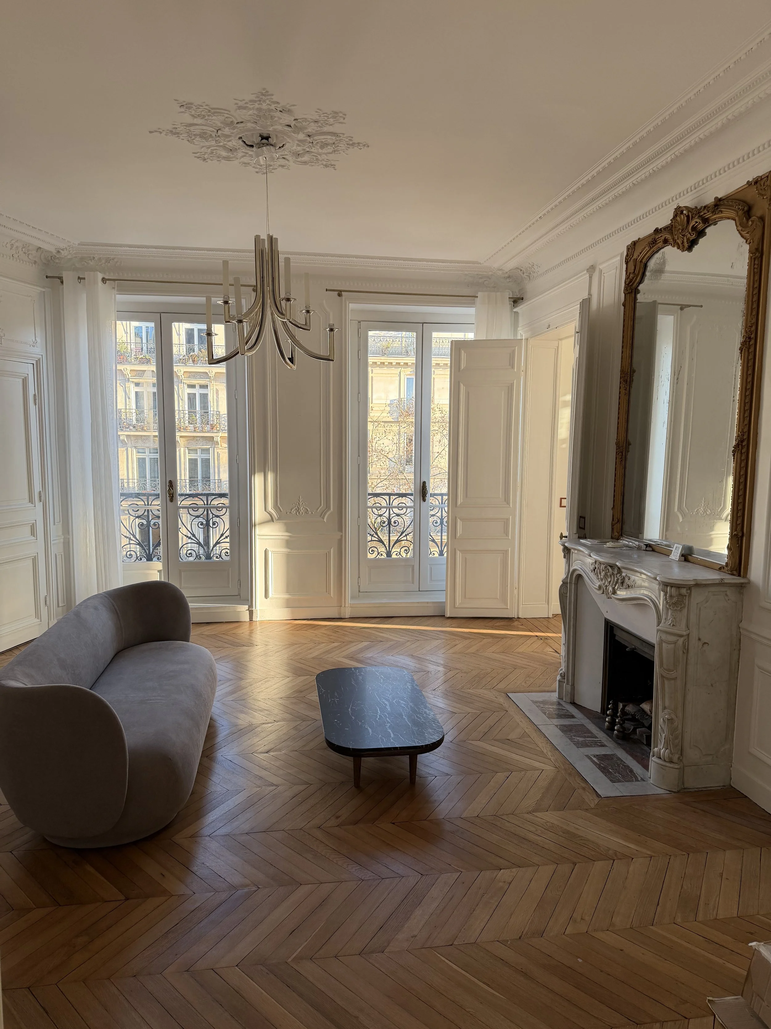 Salon élégant avec parquet en bois, cheminée en marbre, grand miroir doré, chandelier moderne, grands fenêtres avec rideaux blancs, mobilier minimaliste.