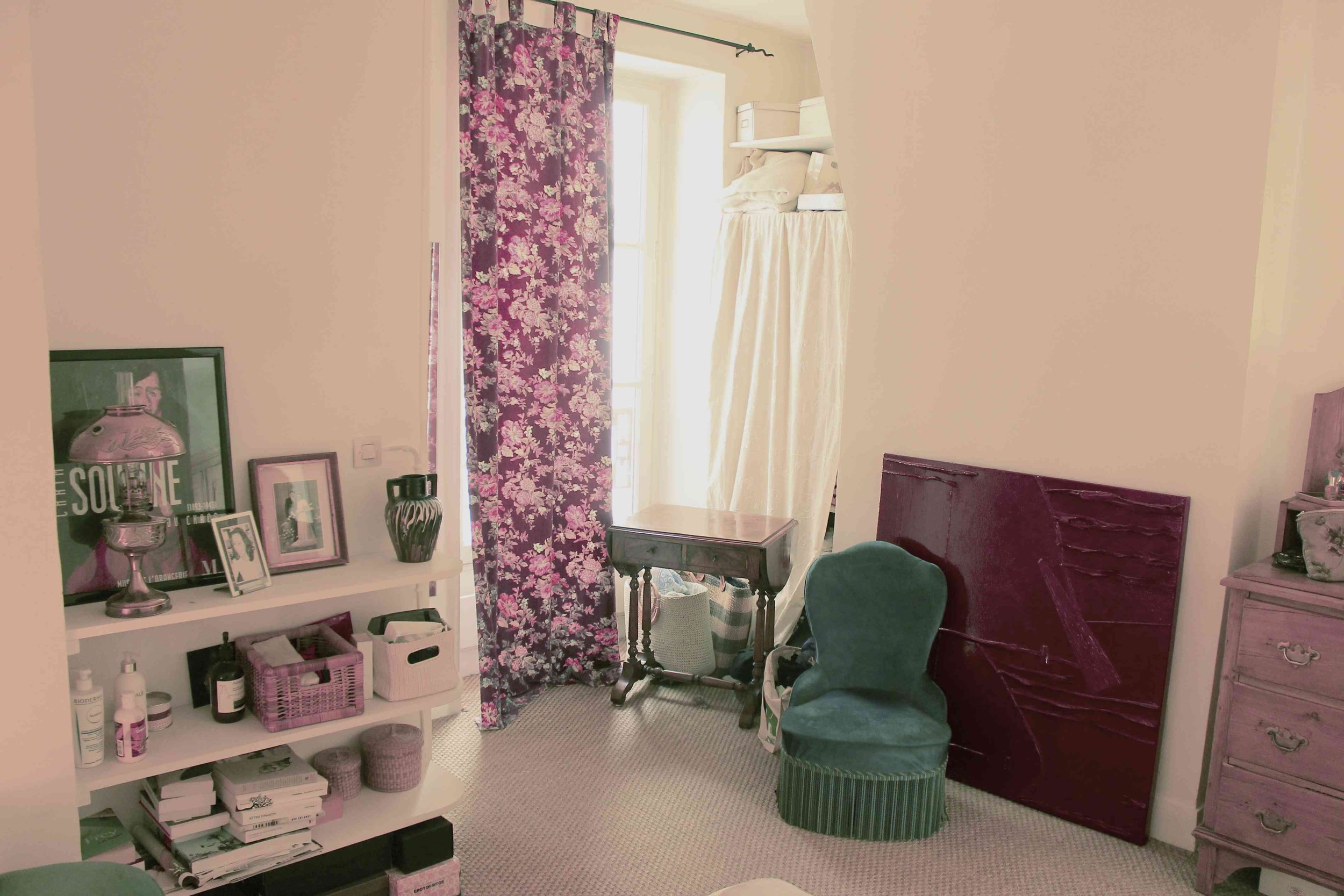Une chambre avec un mur blanc, un tapis beige au sol, une étagère blanche avec des décorations, un grand fenêtre avec des rideaux violets à fleurs, un petit bureau en bois, une chaise verte, une toile ou un cadre rouge, et d'autres meubles et objets décoratifs.