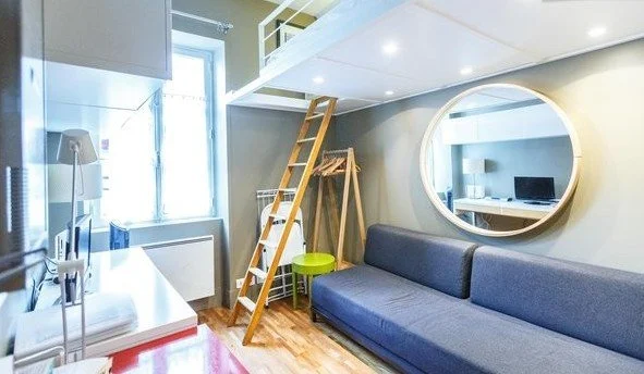Un salon lumineux avec un canapé gris, un mur avec un grand miroir rond, une échelle menant à un espace supérieur, et une fenêtre laissant entrer la lumière naturelle.