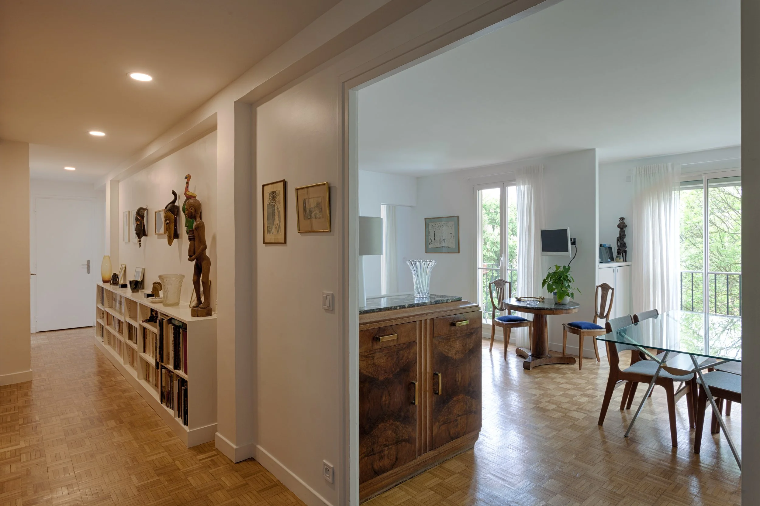 Intérieur d'un appartement avec un couloir à gauche et un salon lumineux à droite, comprenant une table ronde, des chaises en bois, des fenêtres avec rideaux blancs et des décorations murales.