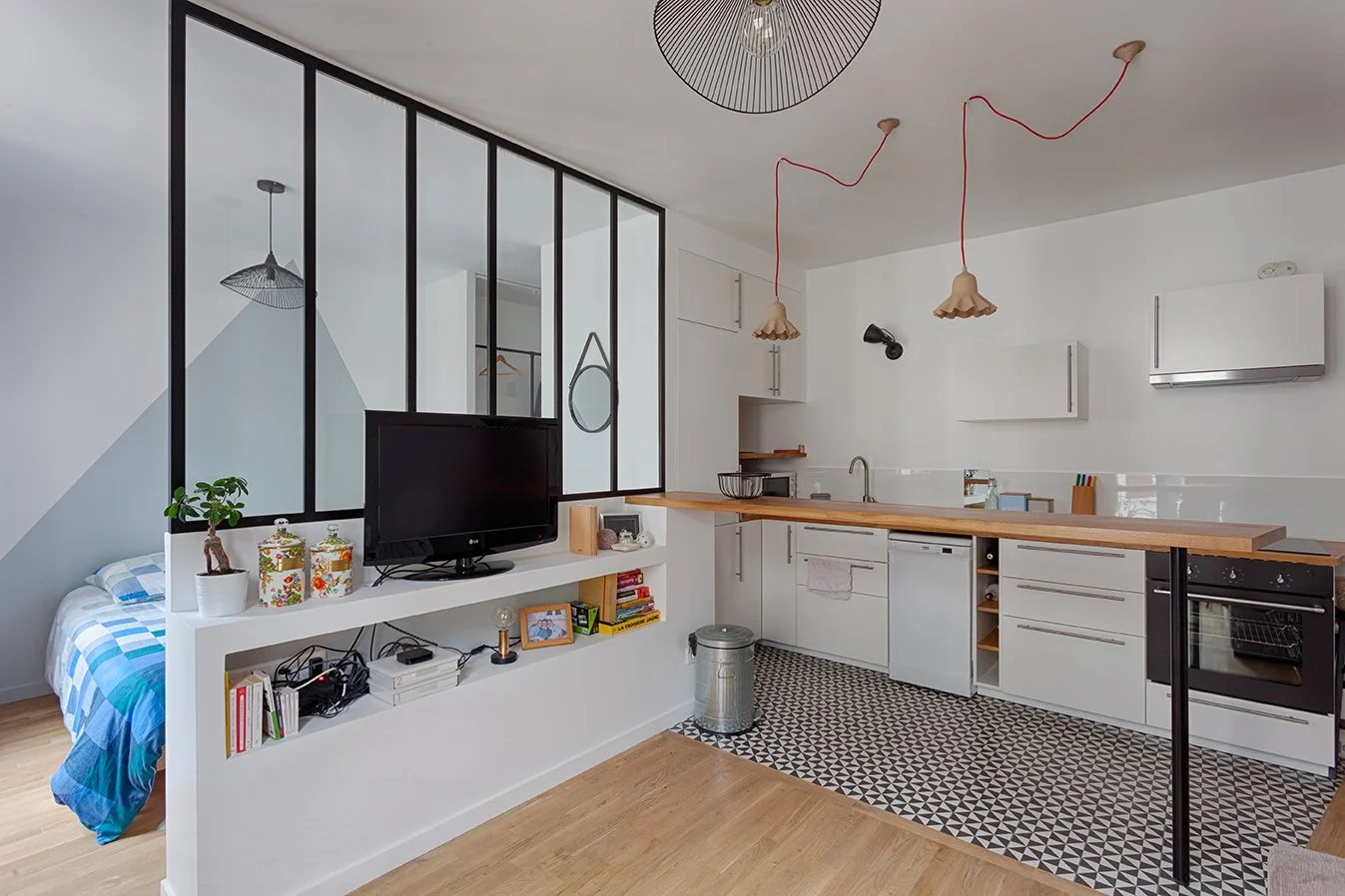 Un studio avec cuisine ouverte, table en bois, étagère avec livres, télévision, et une chambre partiellement visible avec un lit en draps bleus et blancs.