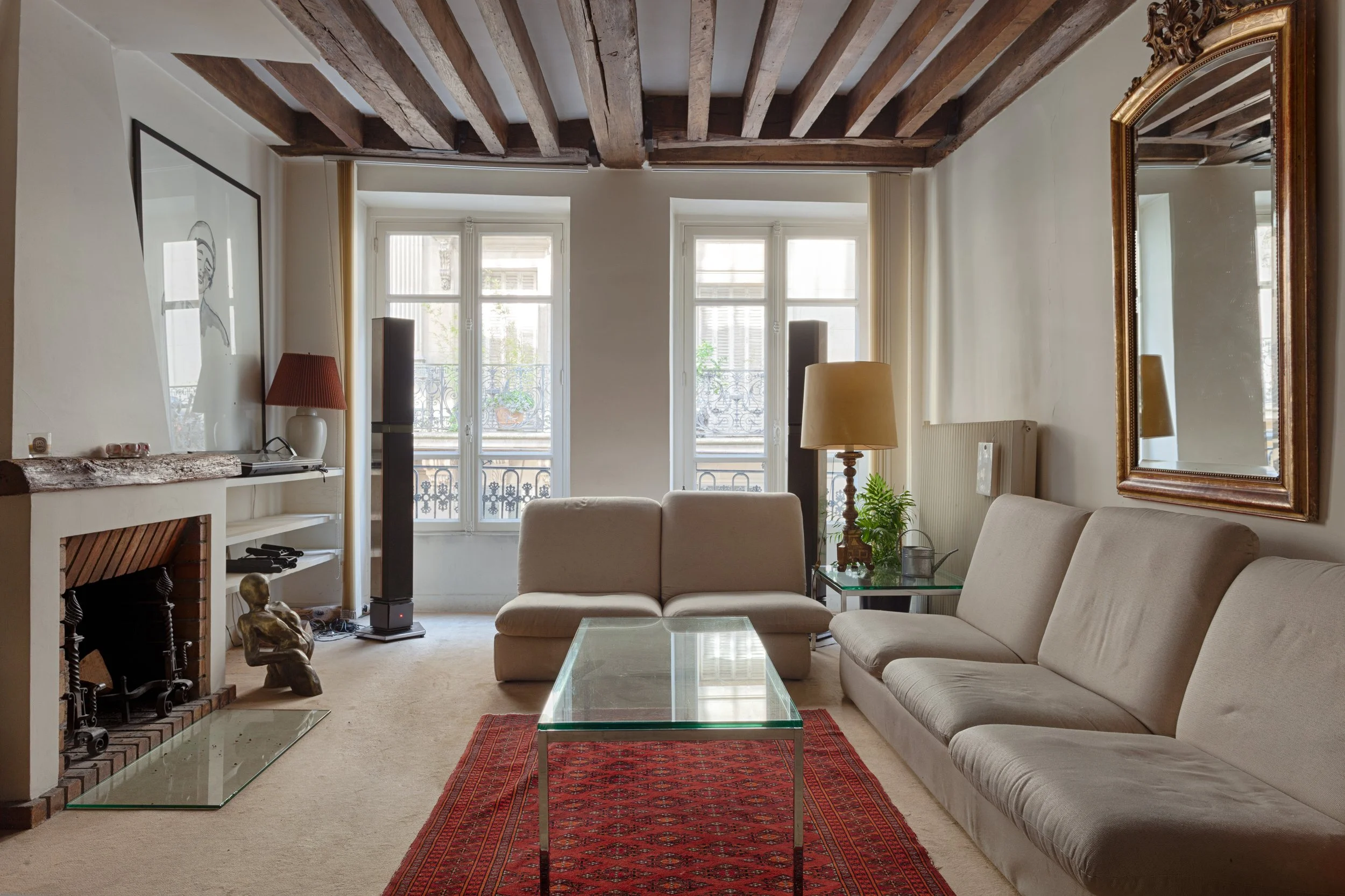 Salon avec deux grands sofas beige, un tapis rouge, une cheminée en briques, une table en verre, des lampes, et des fenêtres laissant entrer la lumière naturelle.