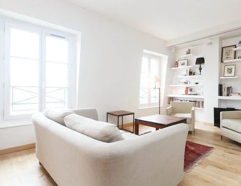Salon moderne avec canapé blanc, fenêtres lumineuses, étagères blanches avec livres et décorations.