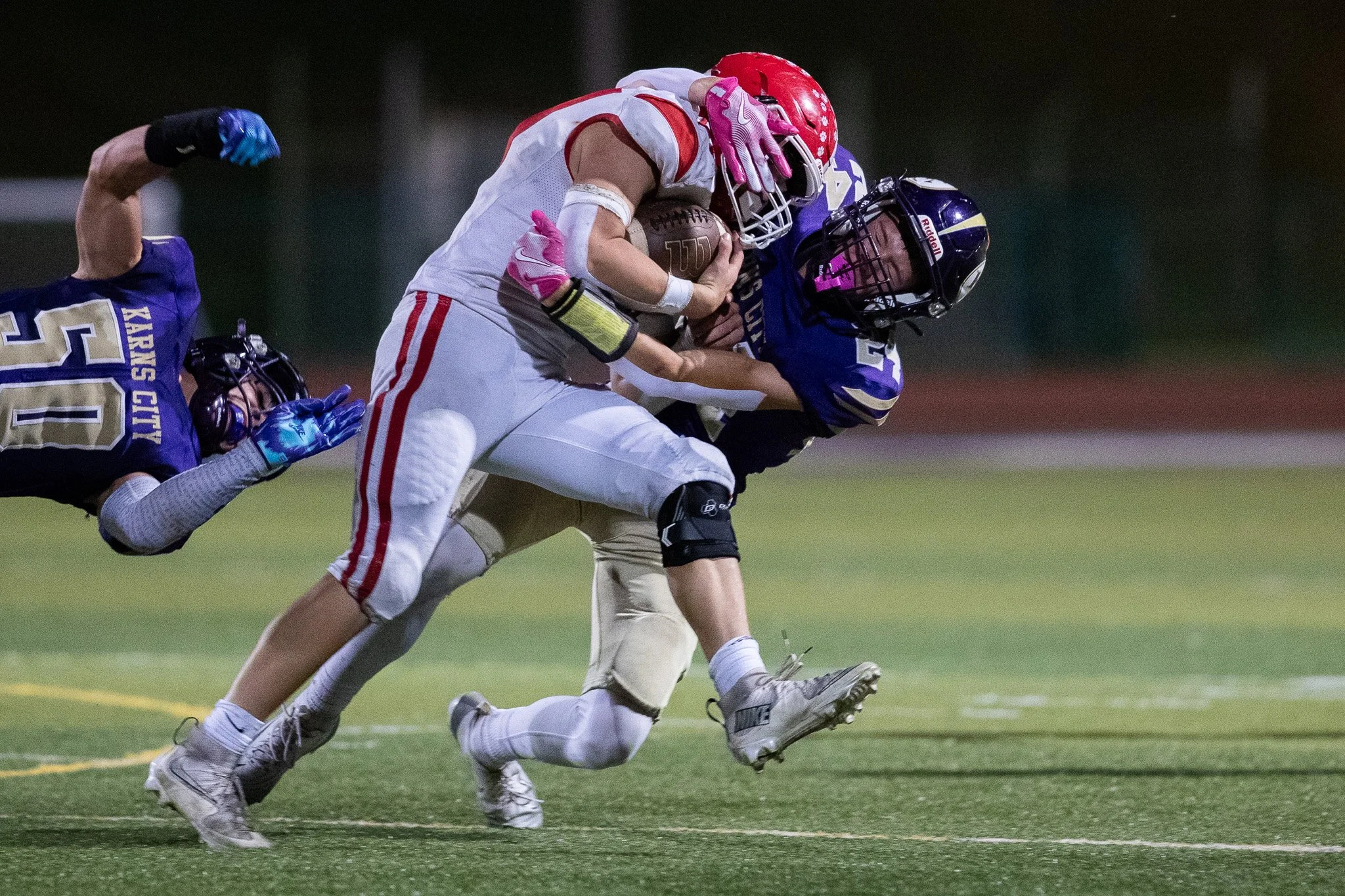 Football players in a tackle during a game, with one player in white holding the ball while being tackled by a player in blue. An opponent in a purple uniform is on the ground nearby.