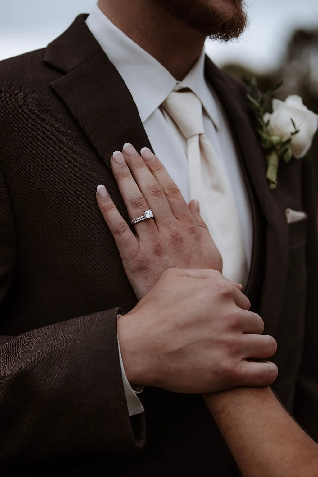 A close-up of a man in a tuxedo with a white shirt and bow tie, wearing a white rose boutonniere, showing his left hand with a wedding ring as a person touches his arm.