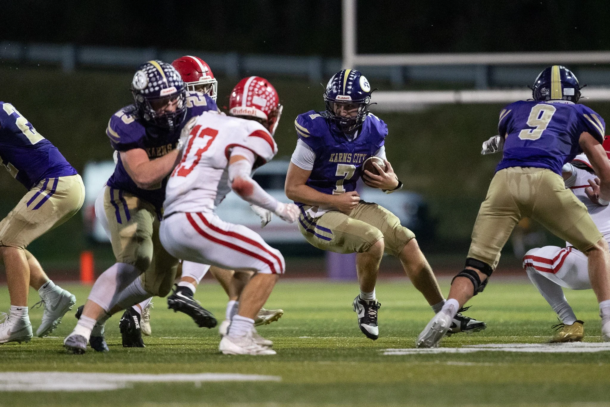 High school football game with players from Karis City in blue and another team in white and red, with a player in blue carrying the ball and running past opponents.