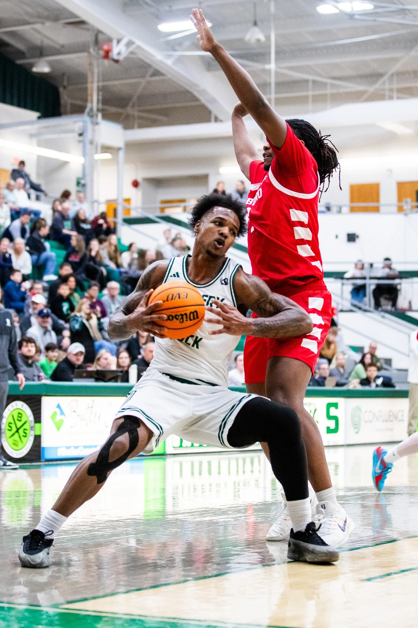 A basketball player in a white jersey is holding a basketball and driving towards the basket while being defended by a player in a red jersey, inside a gymnasium with spectators watching.