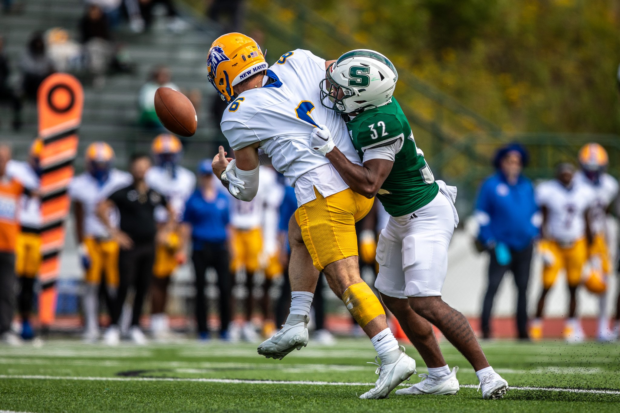 Two football players, one in white and yellow and the other in green and white, collide on the field during a game. The player in white is holding the football, and the player in green is tackling him.