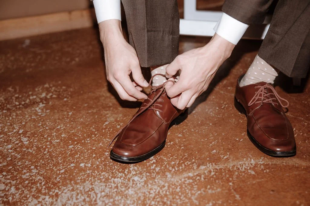 A person in brown dress shoes and polka dot socks is tying their shoelaces, kneeling on a brown textured floor.