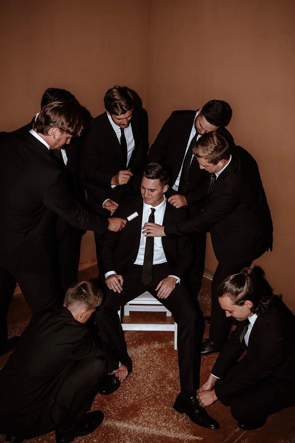 A group of young men in black suits with white shirts and black ties are surrounding a seated man in similar attire, all dressed formally. They appear to be preparing him for a ceremony or event, with some adjusting his shoes and others holding a rol
