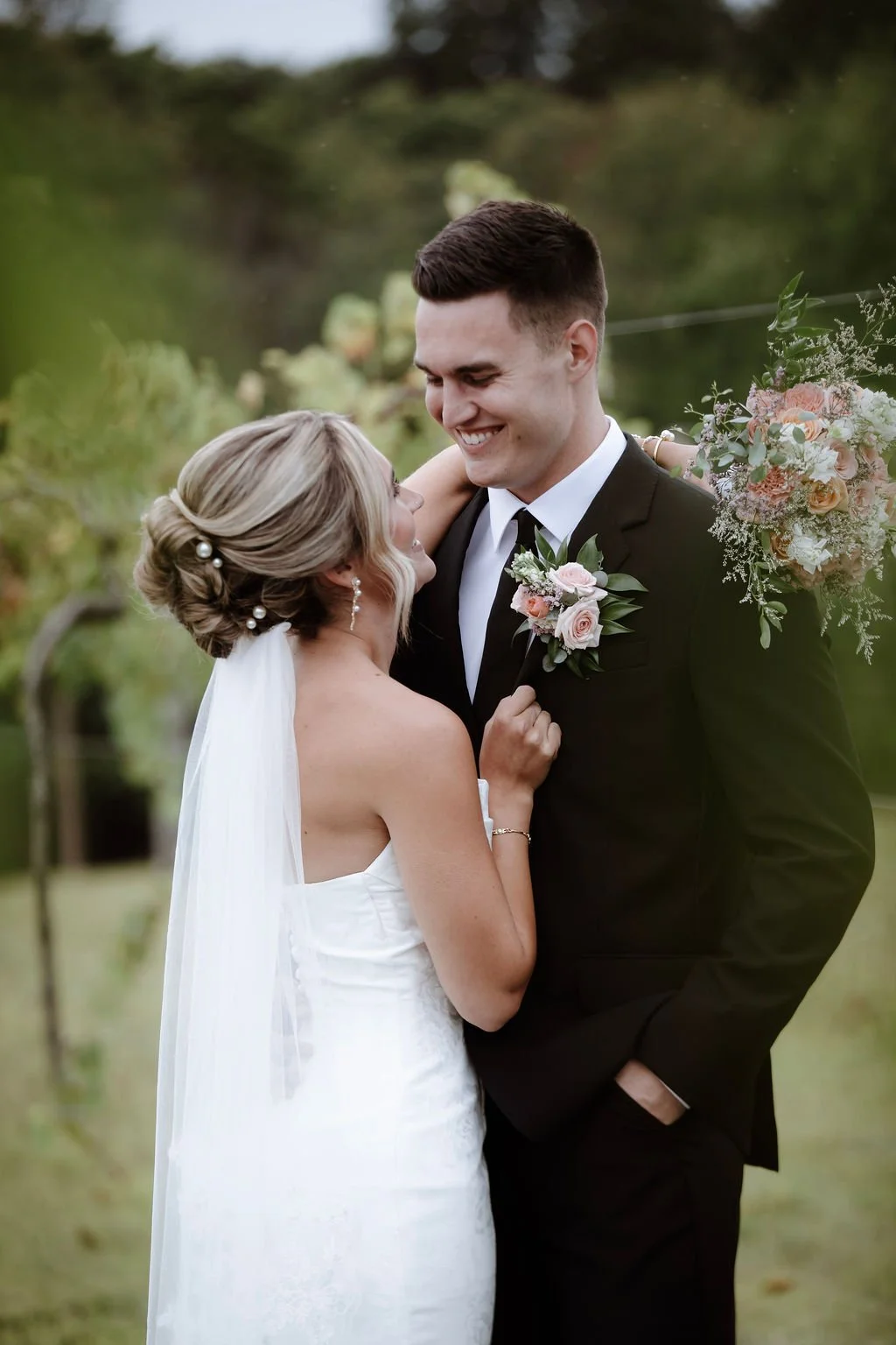 A bride and groom sharing a joyful moment outdoors at their wedding, with the bride in a white strapless dress and the groom in a black tuxedo, holding a bouquet of flowers.