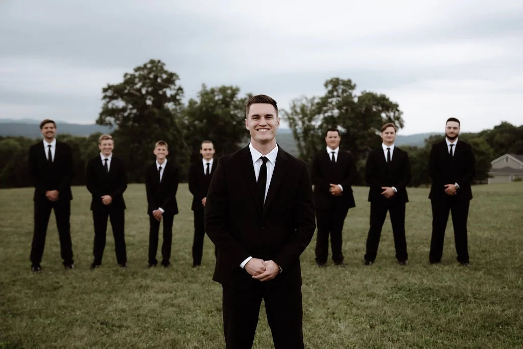 A group of men in black suits and ties standing outdoors on a grassy field, with one man in the foreground smiling directly at the camera and the others standing behind him in a line.