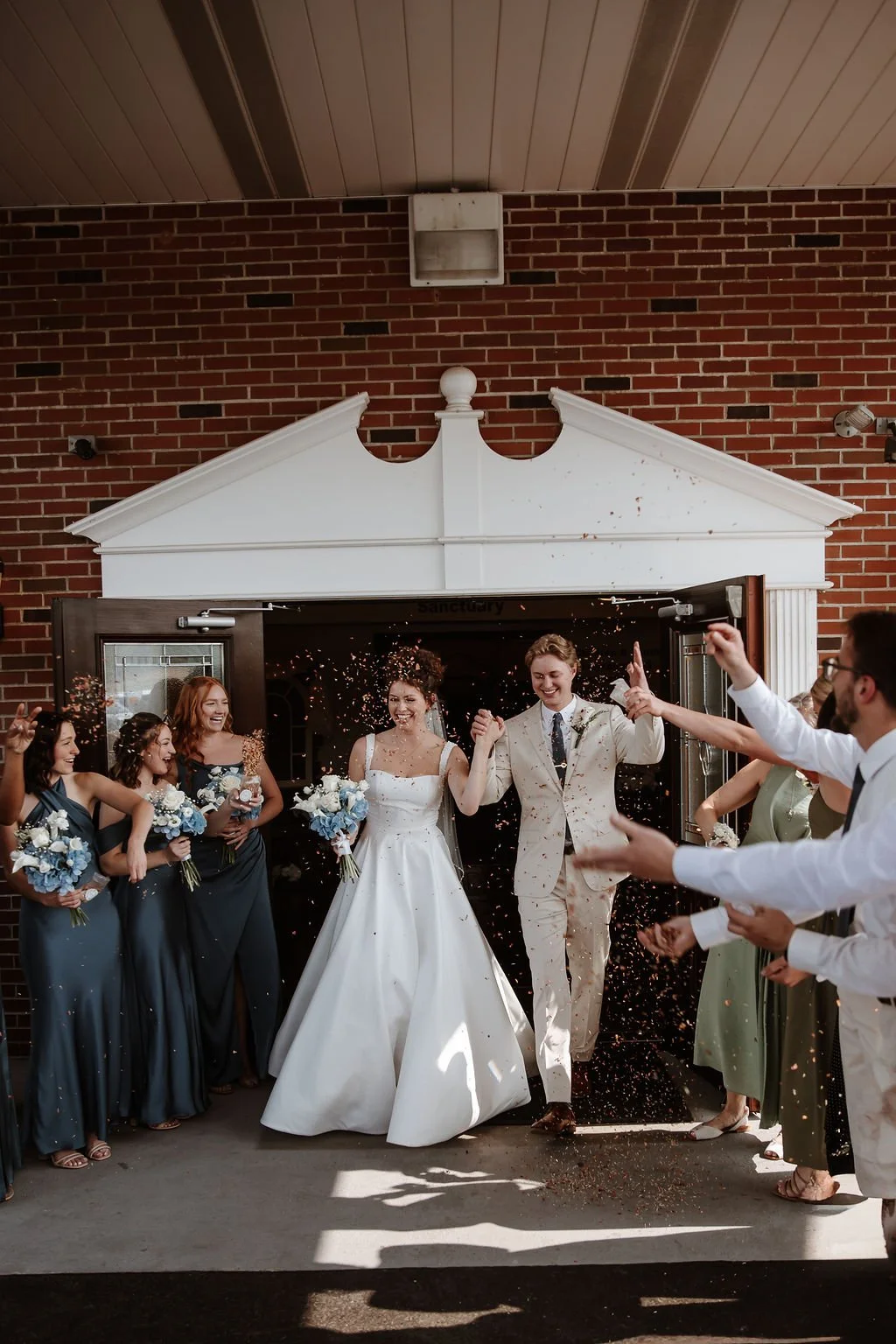 Bride and groom exiting a building with friends and family celebrating outside, some holding bouquets, confetti falling, joyful expressions.