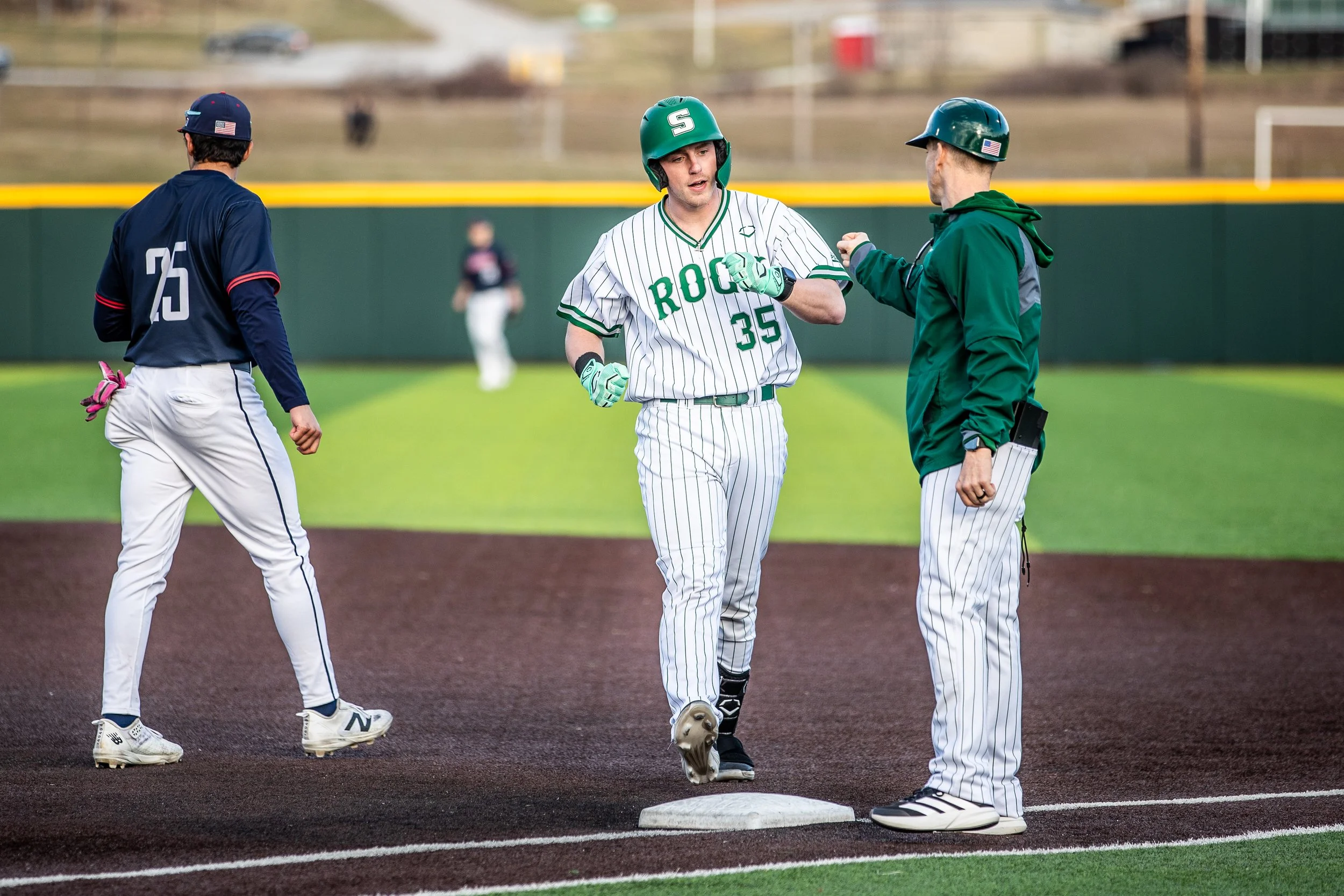 A baseball player wearing a green and white uniform with the number 35 is running on a base while a coach or umpire in green jacket and helmet gives a fist bump. Another player in navy blue and white uniform is nearby. The baseball field has green gr