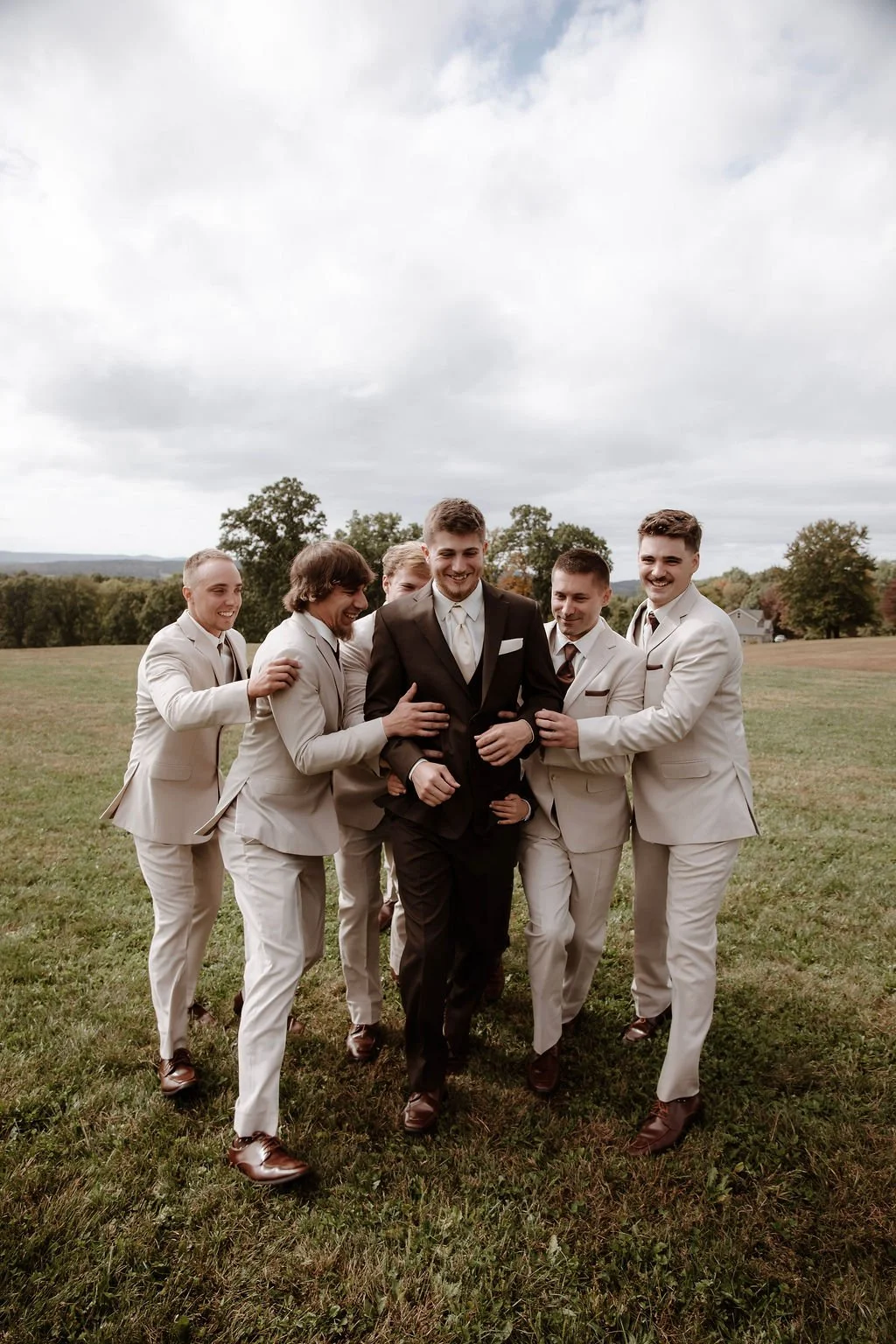 Group of six men, dressed in formal suits, standing on a grassy field under cloudy sky, smiling and enjoying camaraderie, with trees and distant houses in the background.