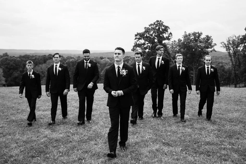 A group of eight men dressed in black suits, walking outdoors on a grassy field with trees and hills in the background, on a cloudy day.