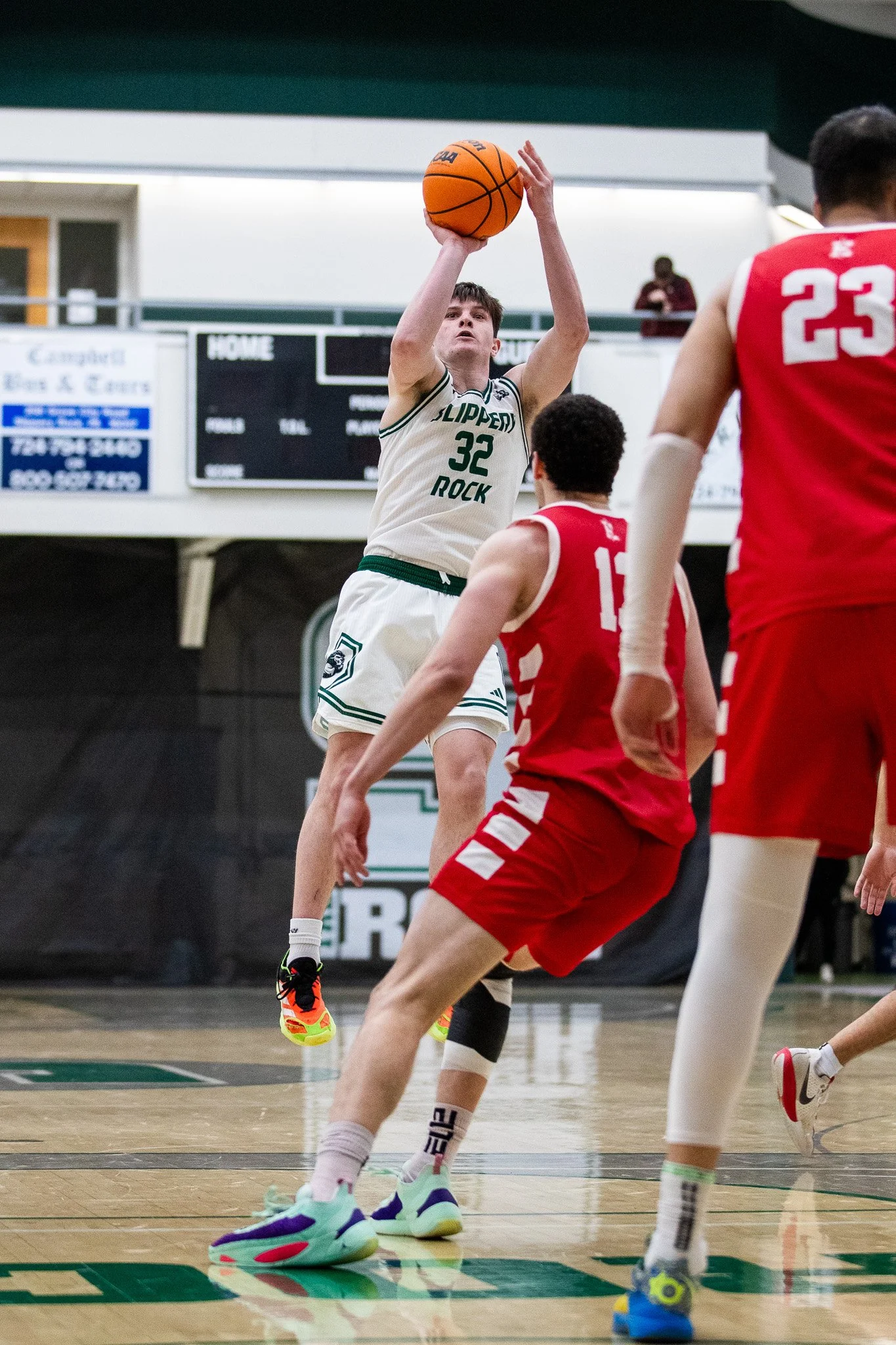 A basketball player in a white jersey with the number 32, jumping and shooting the ball while two opponents in red jerseys attempt to defend.