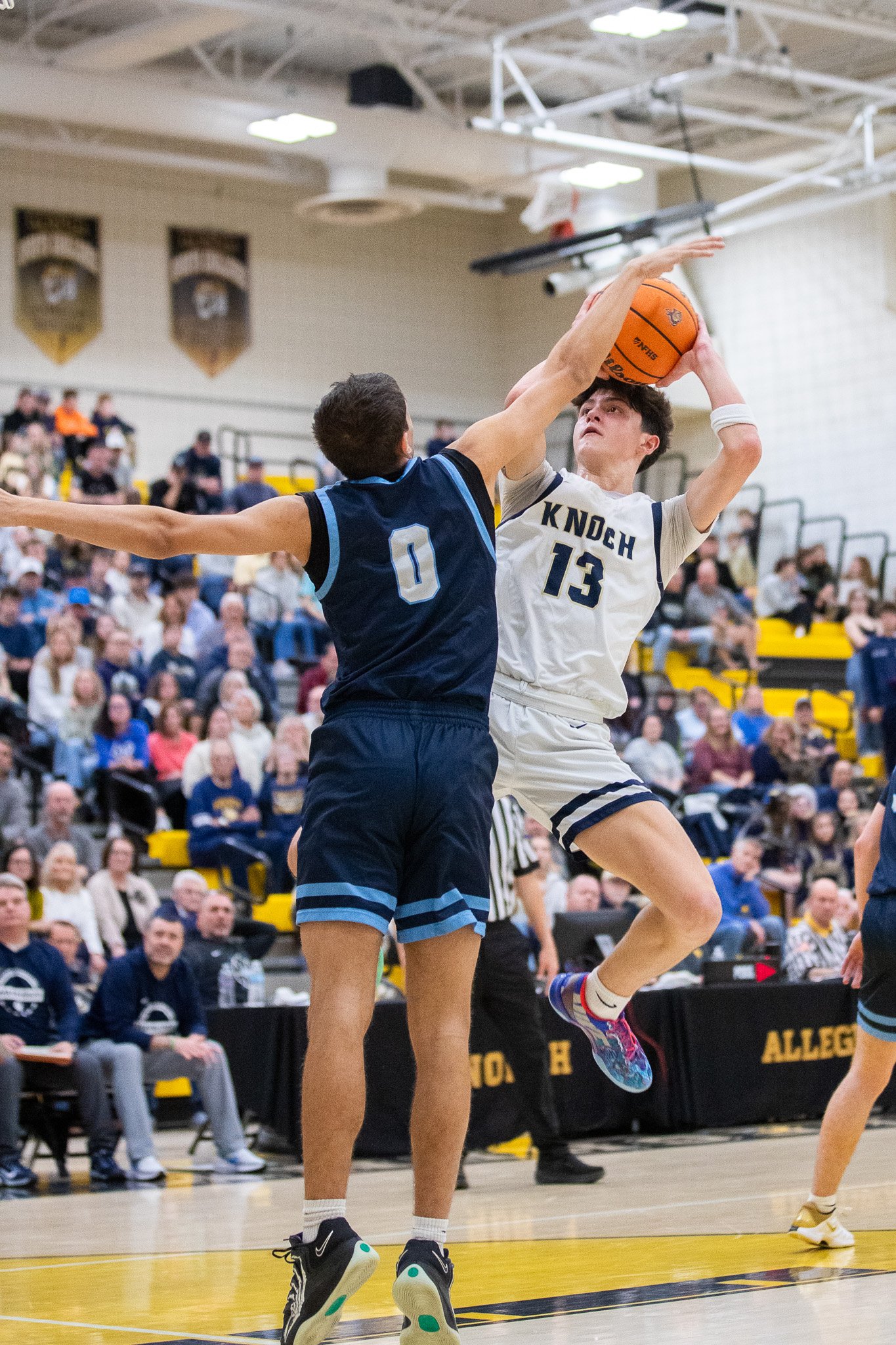 Two basketball players in action during a game, with one player jumping and holding the ball while the other tries to block, in a gym with spectators in the background.