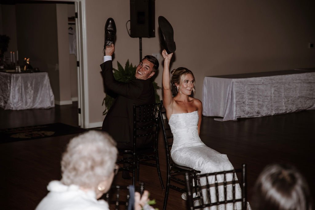Bride and groom participating in a chair game at their wedding reception, each holding shoes in the air while sitting backwards on chairs.
