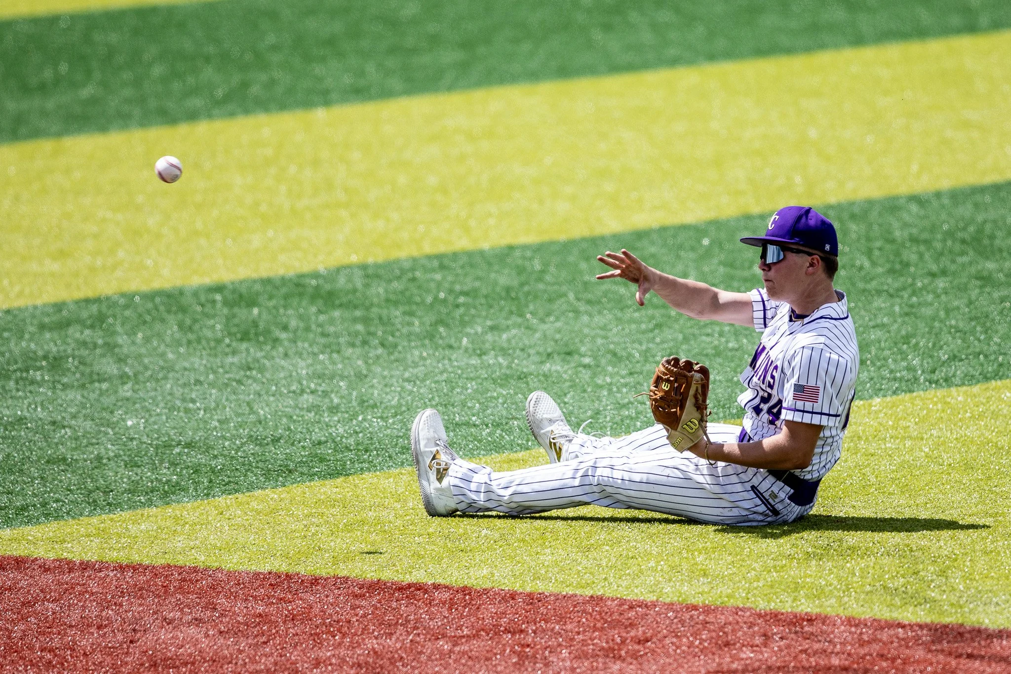 A baseball player in a whitepinstriped uniform, wearing sunglasses and a purple cap, sitting on the field and reaching out with his glove to catch a baseball.