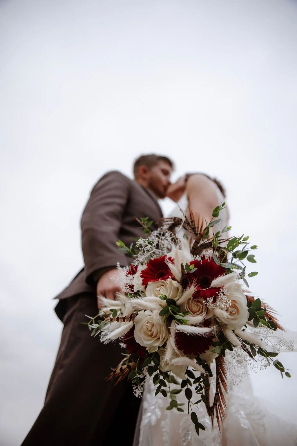 A bride and groom sharing a kiss, with the bride holding a large bouquet of red and white roses and greenery, against a cloudy sky.