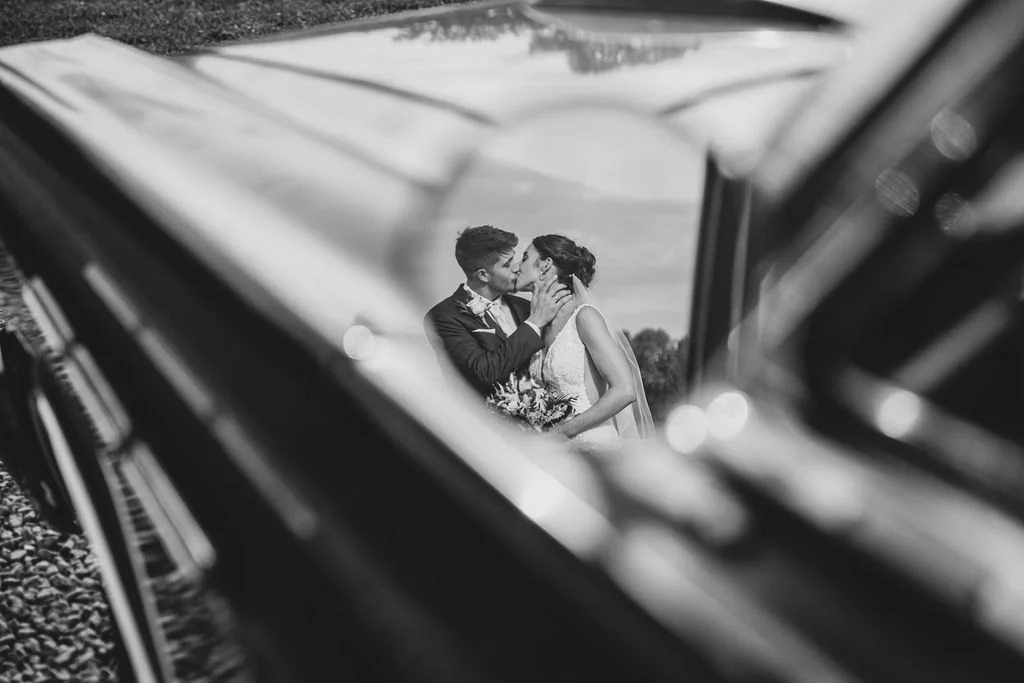 A black and white photo of a couple kissing, viewed through a car window at a distance outdoors.