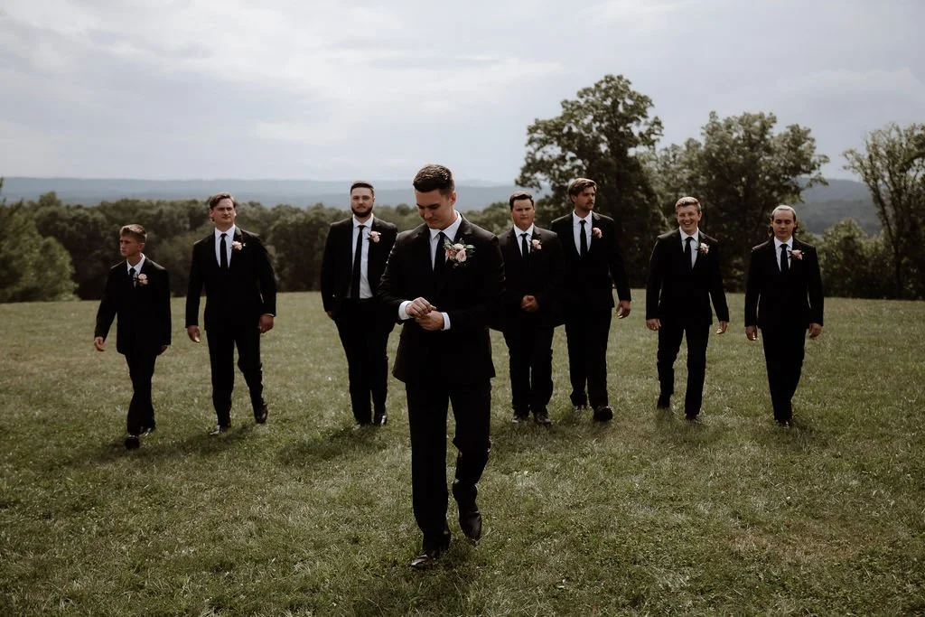 A group of eight men in suits walking across a grassy field, with trees and hills in the background, possibly at a wedding or formal event.
