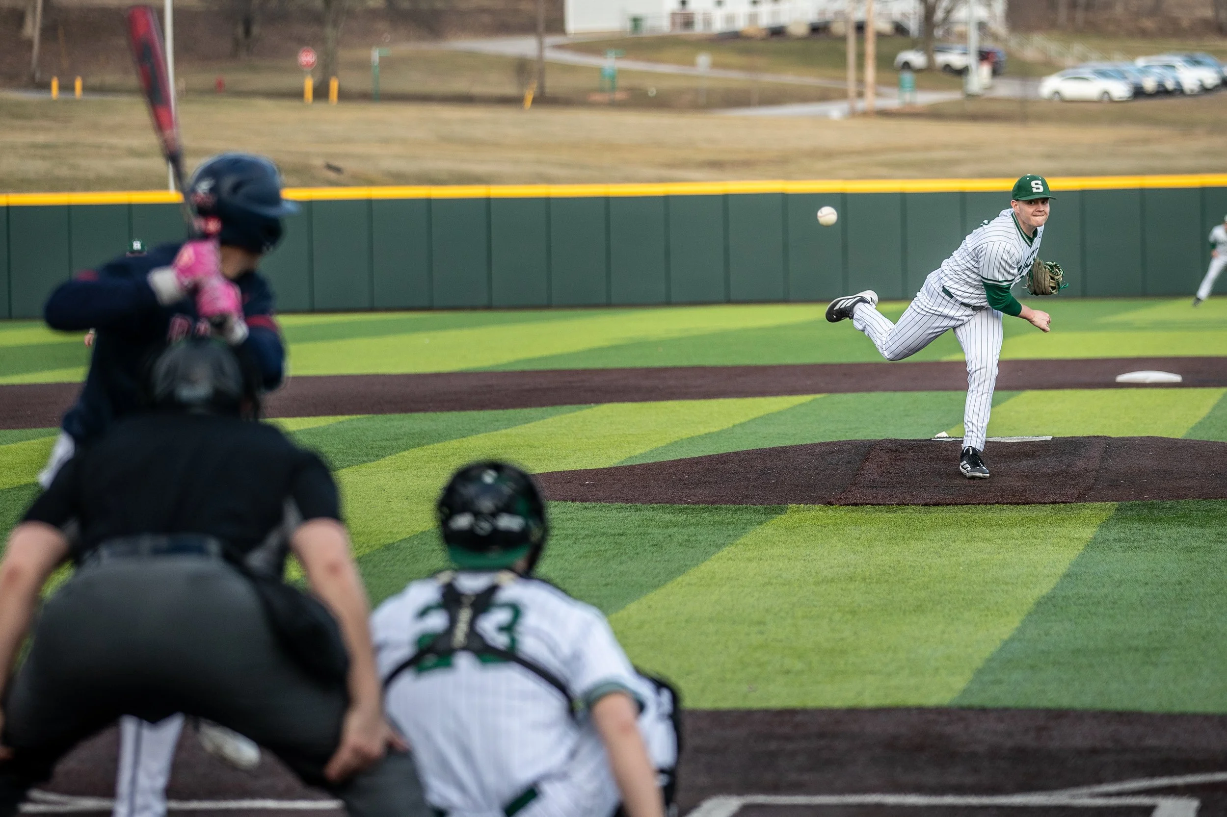 A baseball player in a pinstripe uniform is pitching the ball on the mound while an opposing batter and home plate umpire watch from behind. The scene is on a green baseball field with a dark brown pitcher's mound and outfield fence, with a few cars 