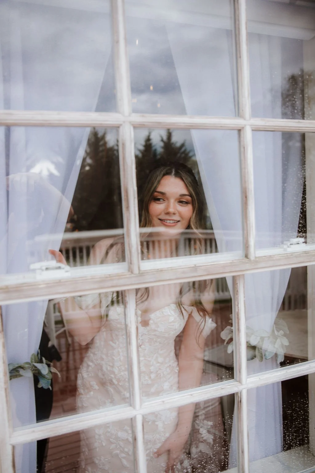 A woman in a wedding dress looking out a window with white curtains, smiling.