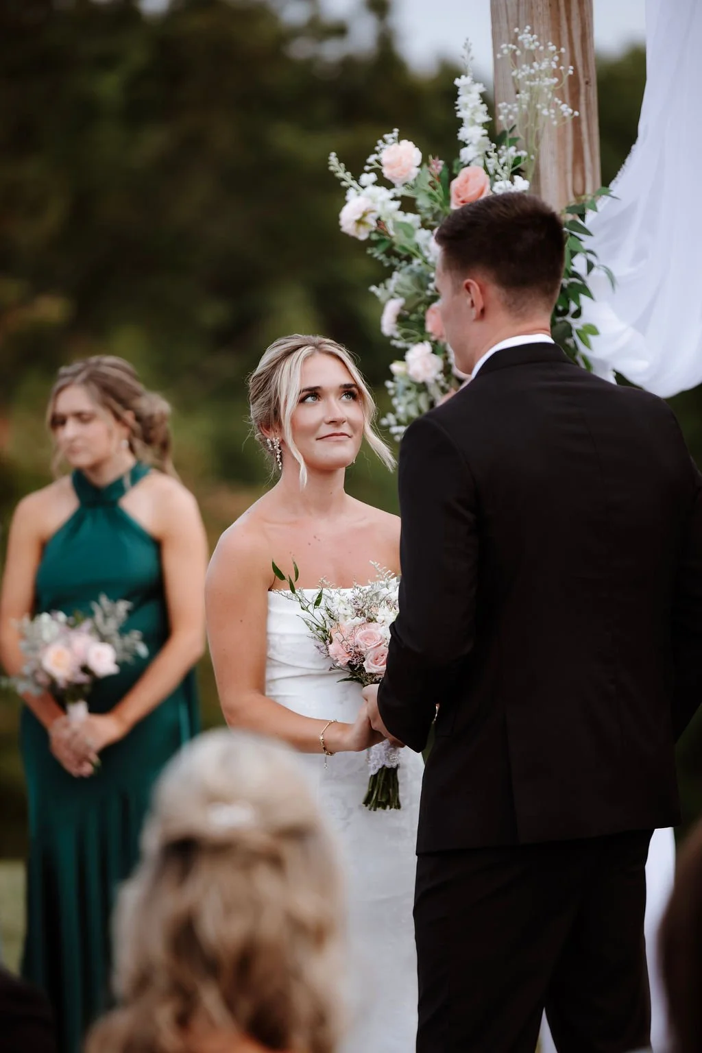 A wedding ceremony outdoors with a bride, groom, and bridesmaid near a floral arch decorated with pink and white flowers. The bride is gazing at the groom, holding a bouquet of flowers, with an emotional expression.