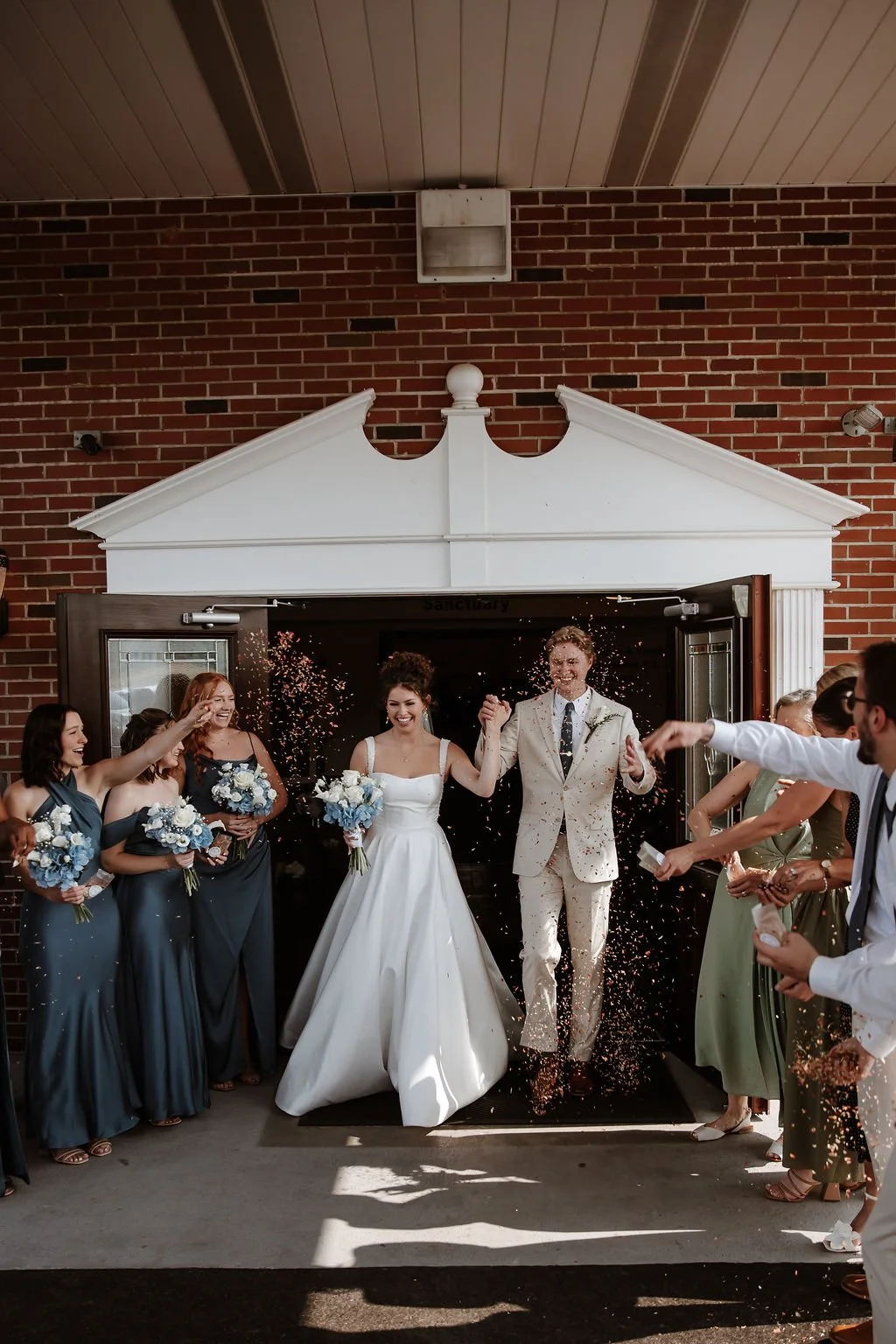 Bride and groom exit a building with friends throwing confetti, celebrating their wedding.