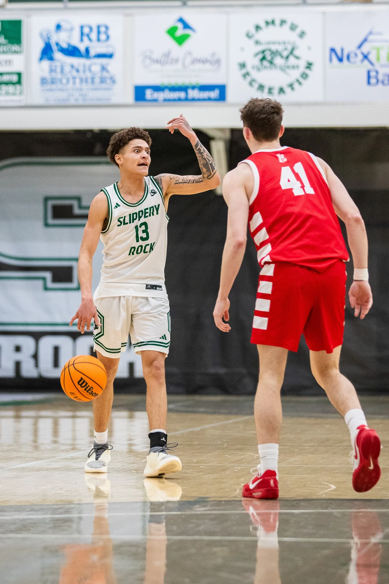 Two basketball players on the court during a game, with one player in a white uniform and the other in a red uniform, having a conversation.