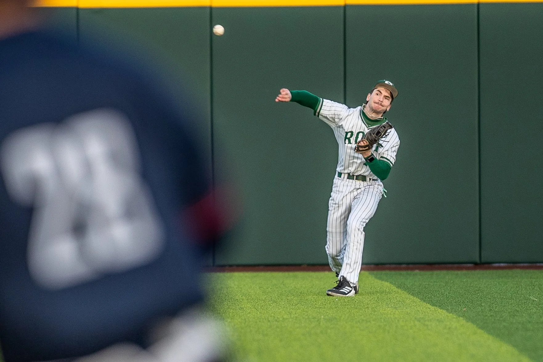 A baseball player in a striped uniform and cap is preparing to catch a ball against a green outfield wall, with a blurred figure in the foreground.