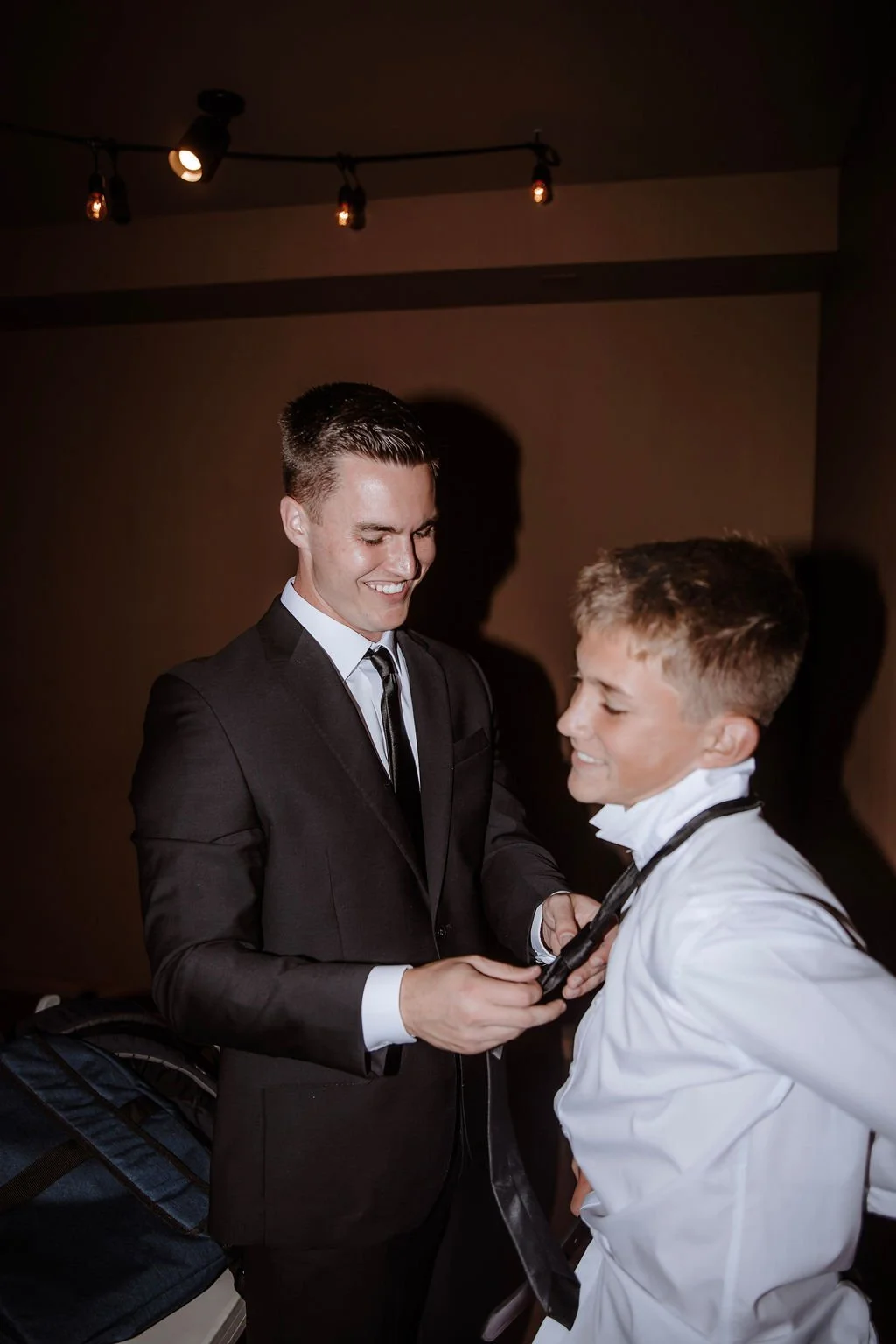 A young man in a black suit and tie is helping another young man adjust his tie, who is dressed in a white shirt with a black bow tie. They are smiling and appear happy.