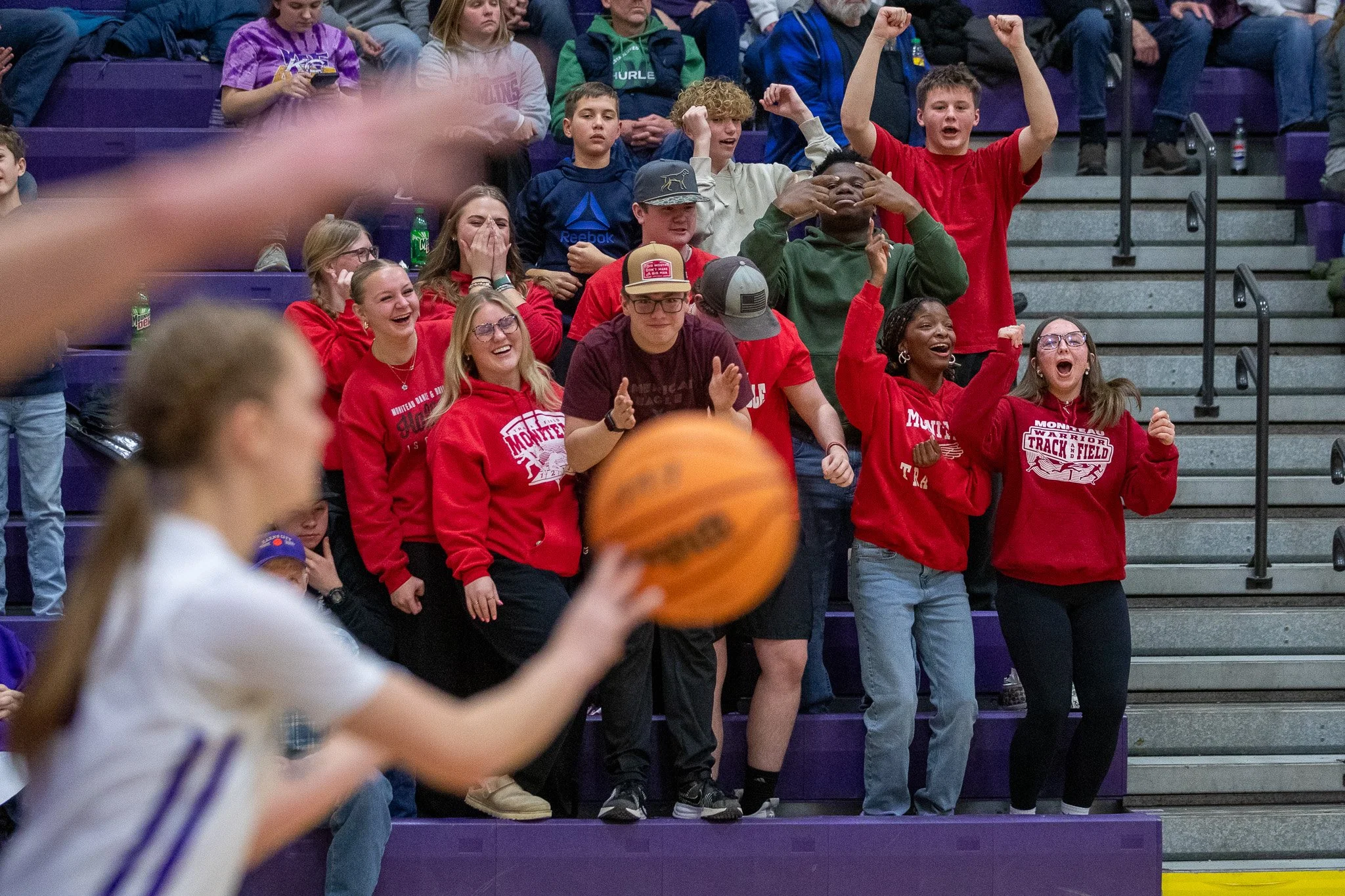 Group of students cheering and clapping during a basketball game at school gymnasium, with a student in the foreground shooting a basketball.
